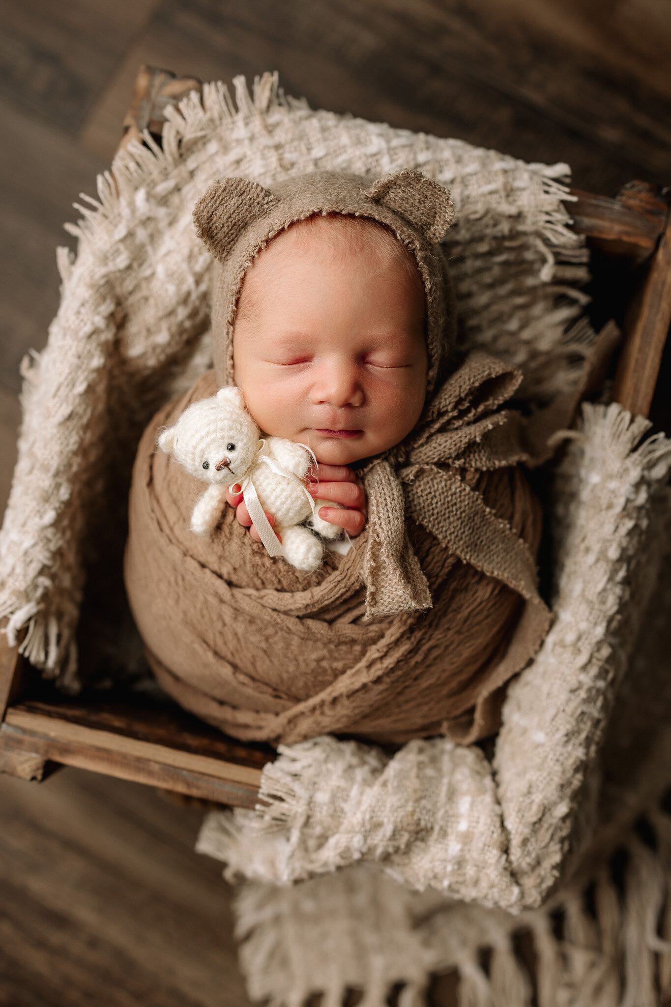 Baby boy swaddled in a brown wrap, wearing a bear bonnet, and snuggling a tiny white teddy bear