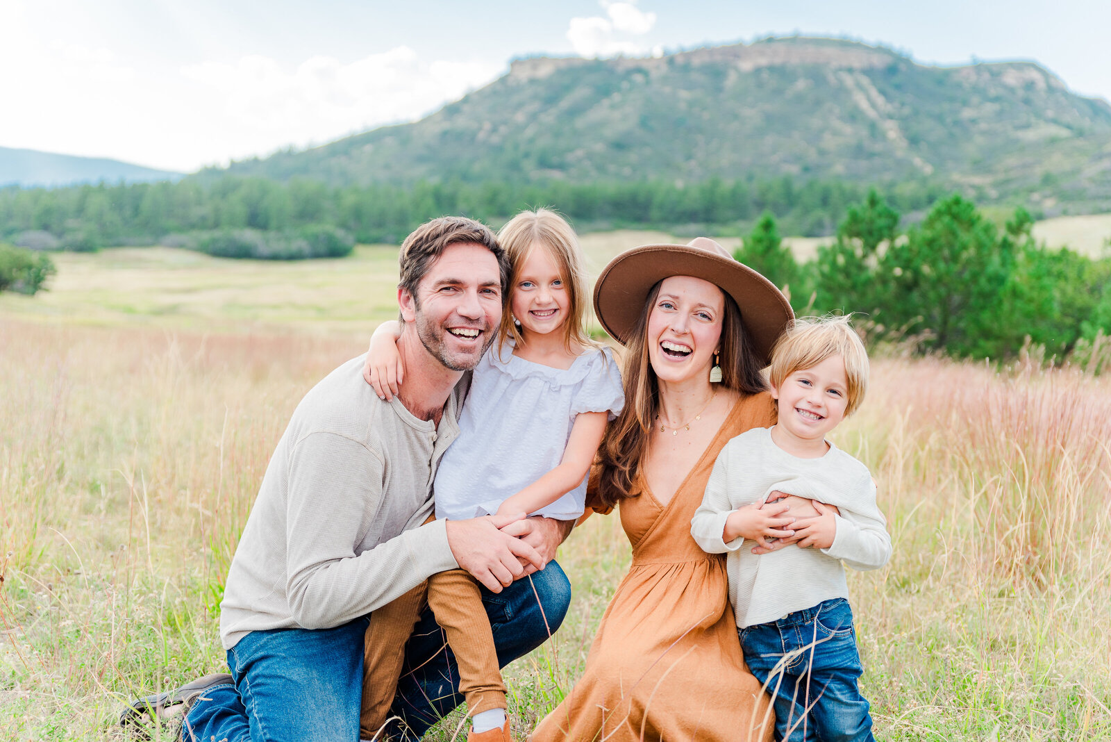 A family with the mom in an orange dress and tan hat, dad in a tan top and jeans and kids both in neutral colored clothing all cuddle close together while squatting in the grassy field capturing by denver family photographer