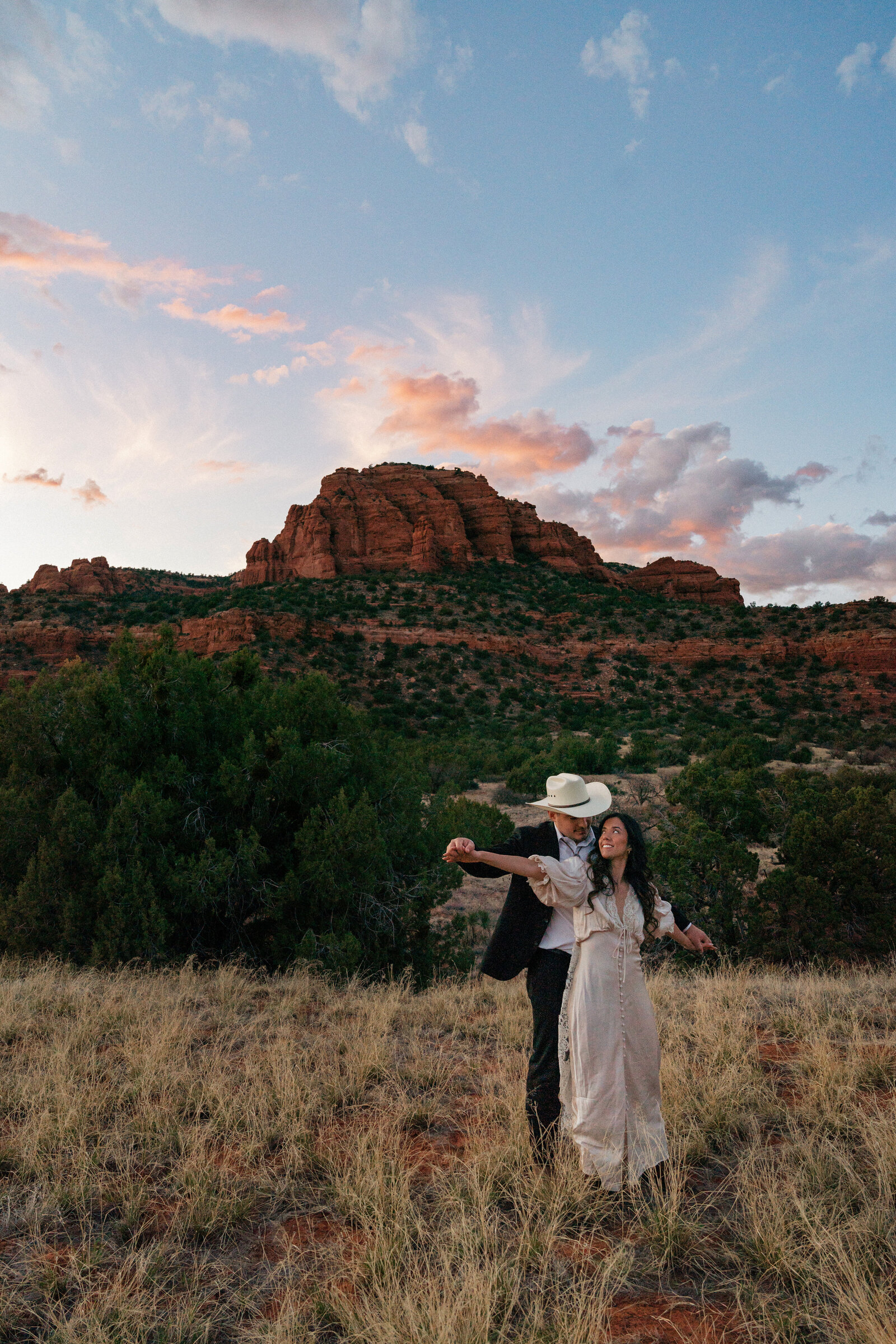 Bride and groom in western attire dancing at sunset during their Bear Mountain elopement in Sedona, surrounded by red rocks and a glowing sky.