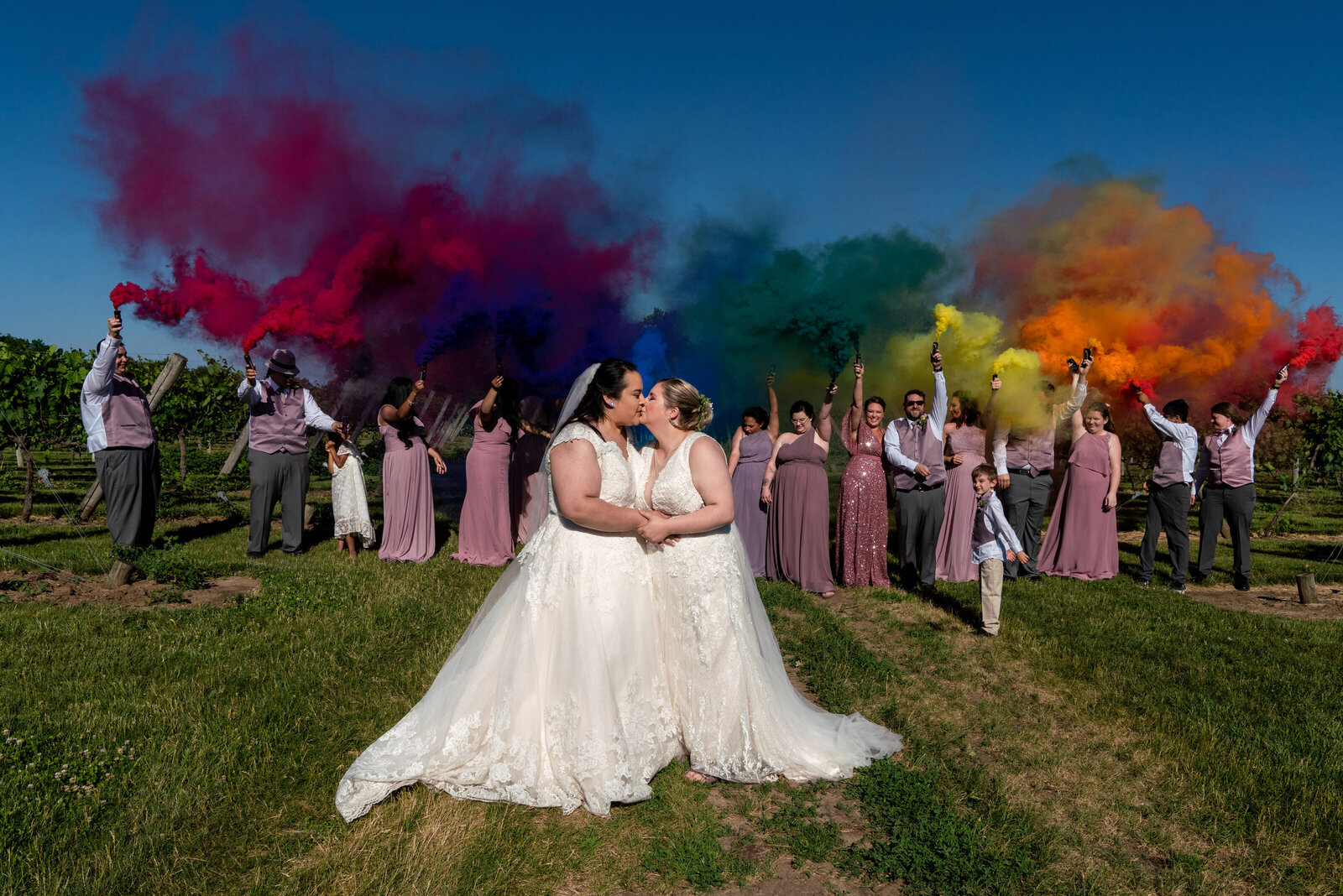 Lesbian brides kiss in front of rainbow colored smoke on their wedding day.