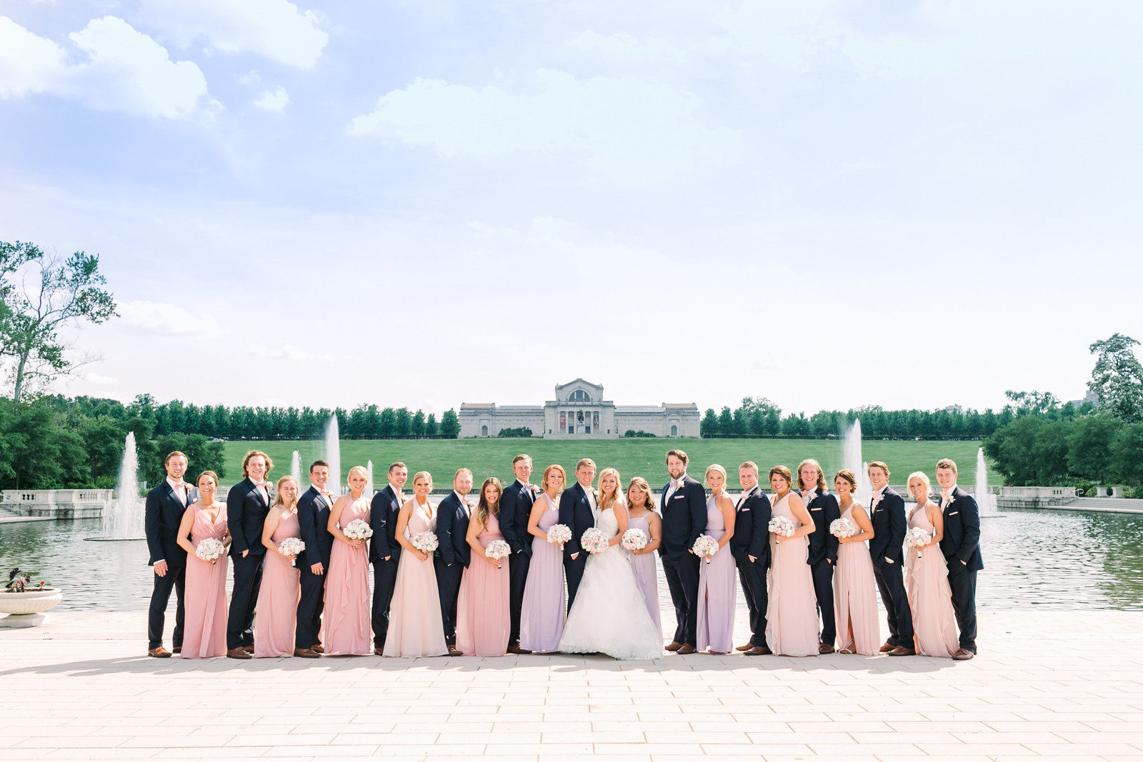 Molly and Marcus's wedding party of 22 people pose in front of the St. Louis Art Museum and Grand Basin with a beautiful clear blue sky.