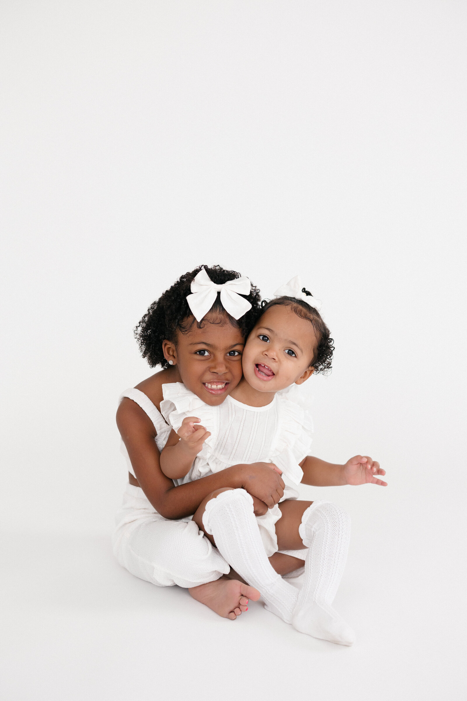 Two sisters snuggling and smiling as they wear all white and sit on a white backdrop