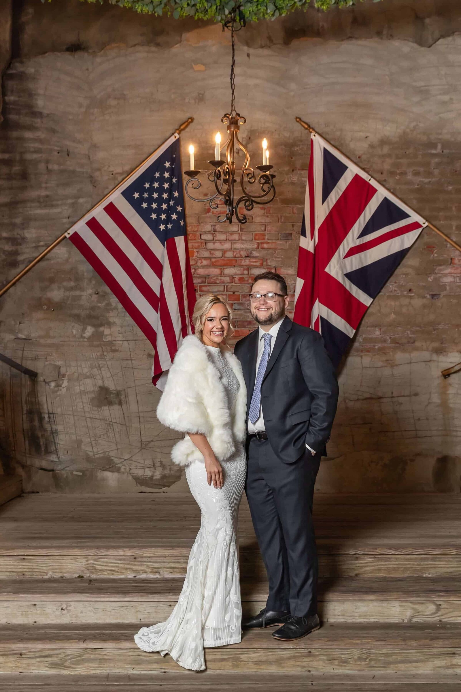 A bride in a white dress and fur shawl stands next to a groom in a dark suit, standing beneath a chandelier between a U.S. flag and a U.K. flag against a brick wall.