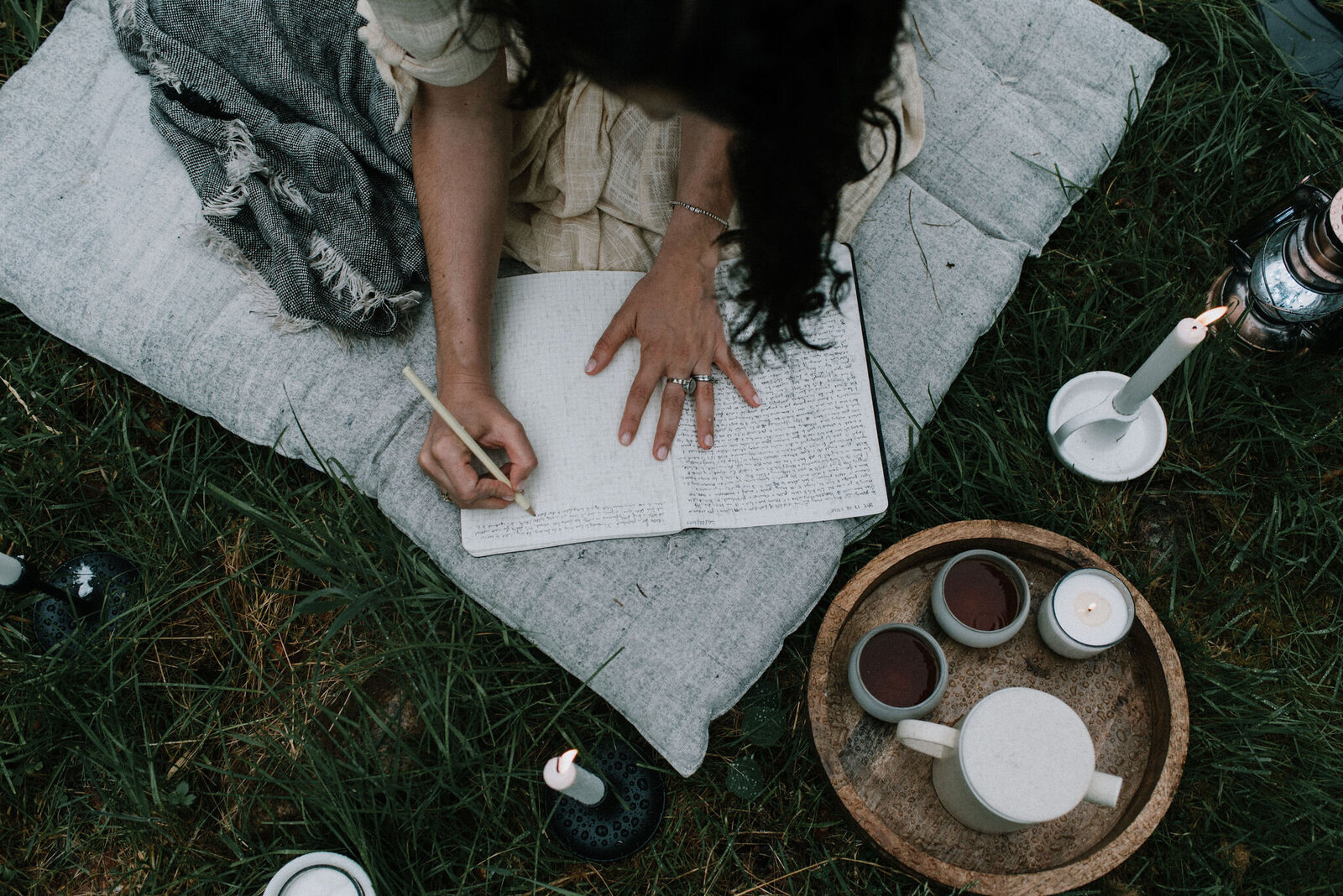 a woman is writing in a journal placed on a pillow on the grass, next to candles and tea, all covered in raindrops