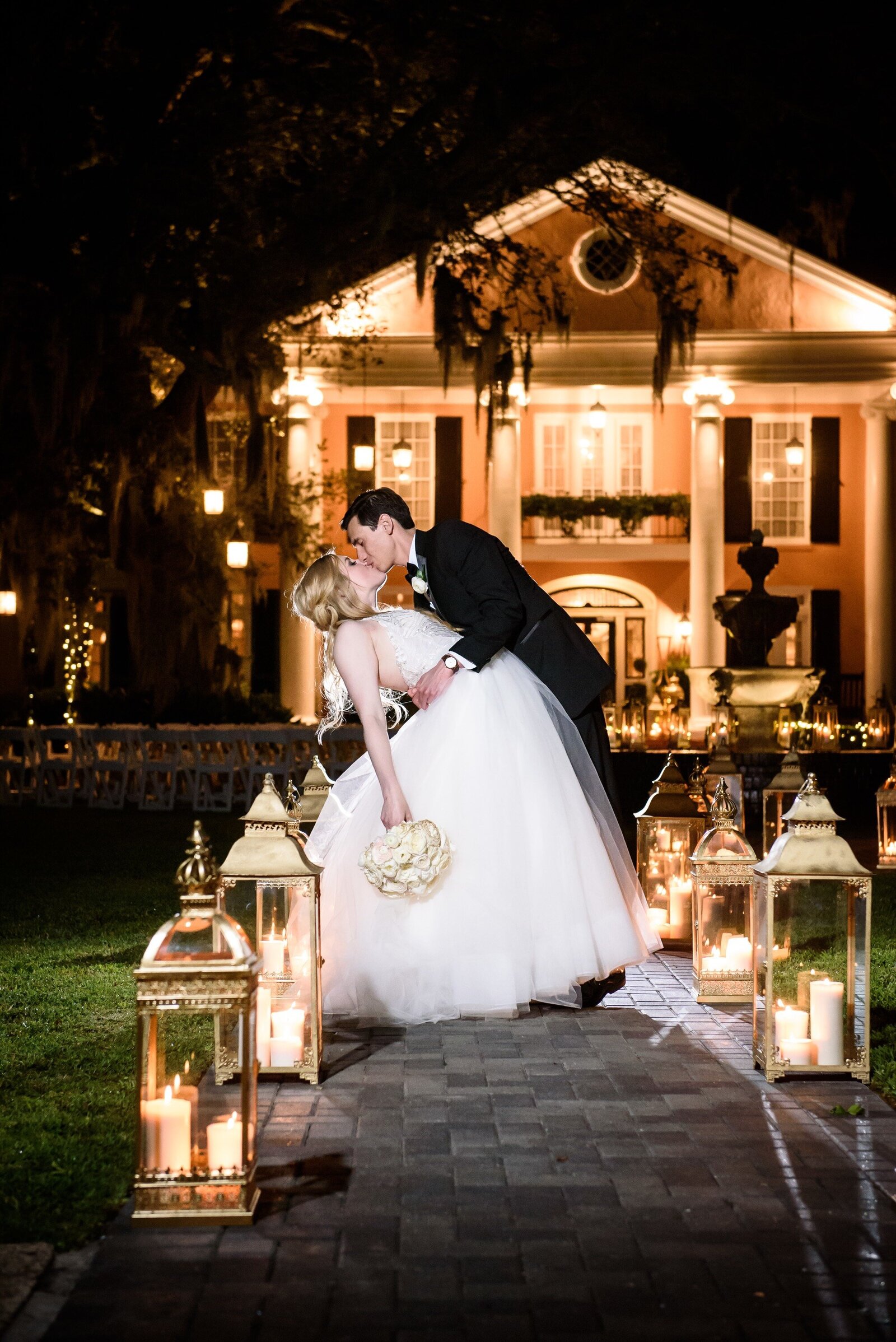 Bride and groom dip and kiss in front of Southern Oaks with lanterns lining the walkway