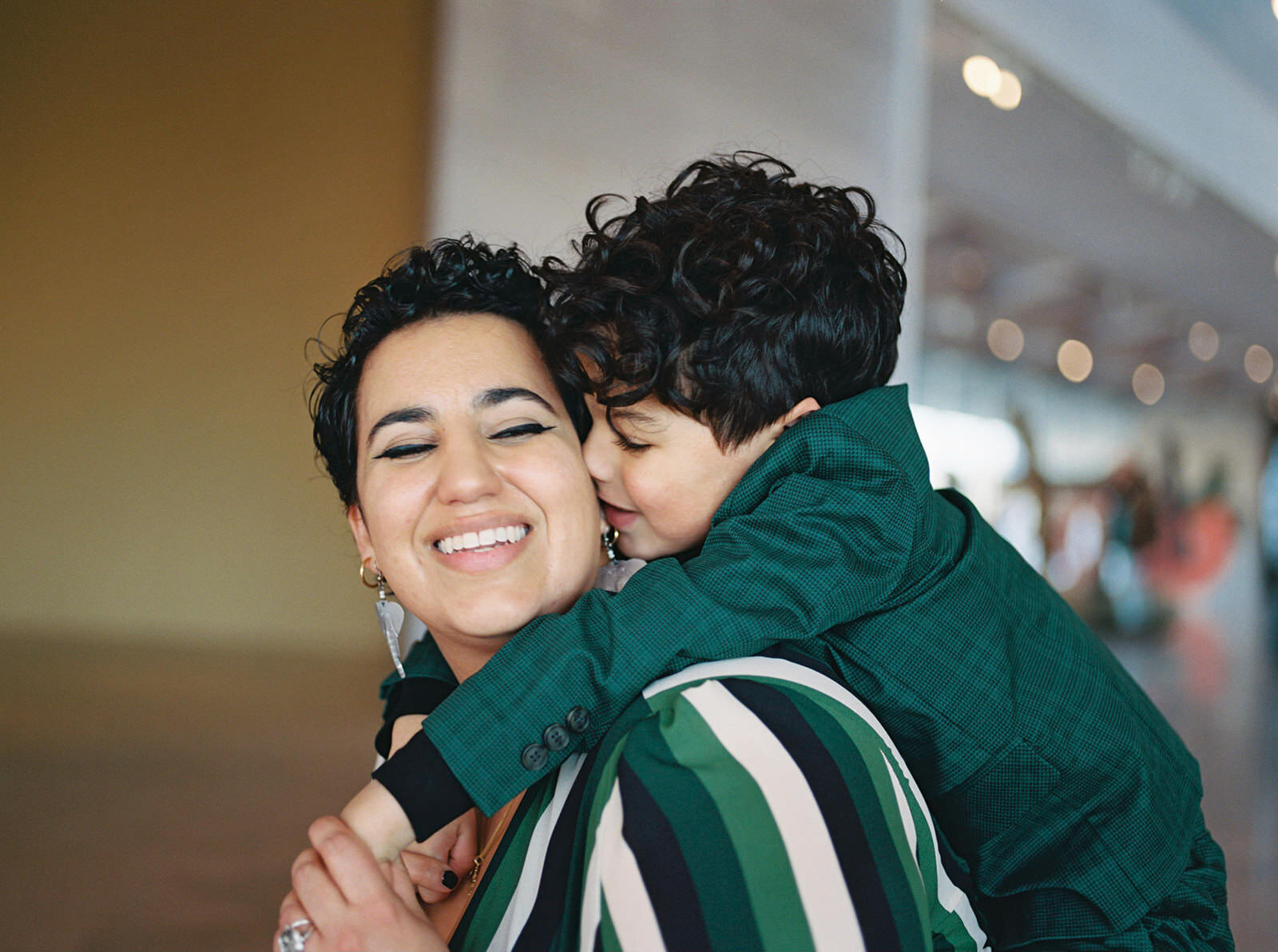 A joyful portrait of a mother and her young son, with the child hugging her tightly from behind, both smiling brightly in a modern indoor setting.