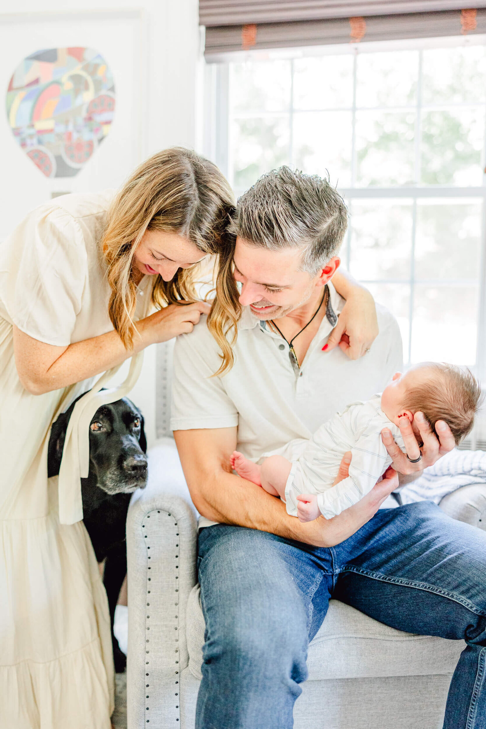 Mom and dad laugh at a black lab who is sneaking under mom's dress to see the newborn baby dad is holding