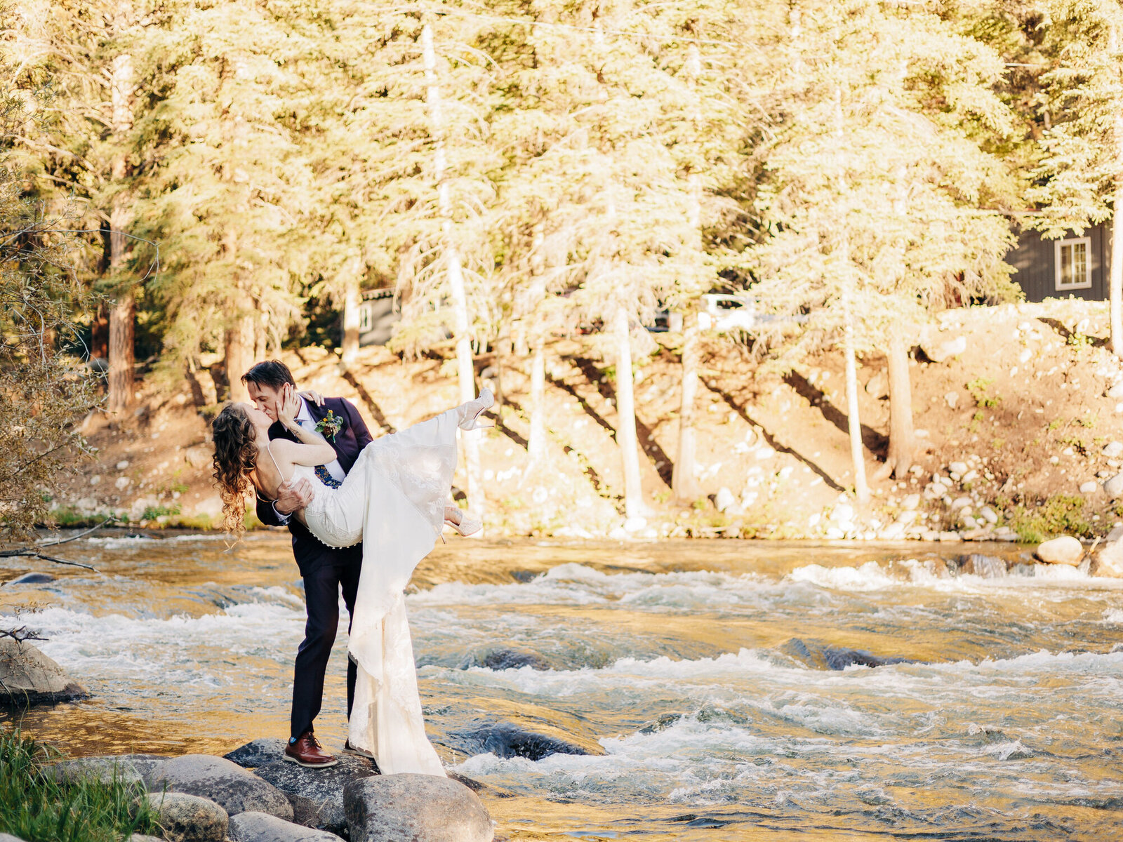A groom lifts and kisses his bride beside a flowing river, surrounded by tall trees and sunlight. Captured by an Eau Claire Wedding Photographer, the bride’s dress trails down as they stand on rocks near the water.