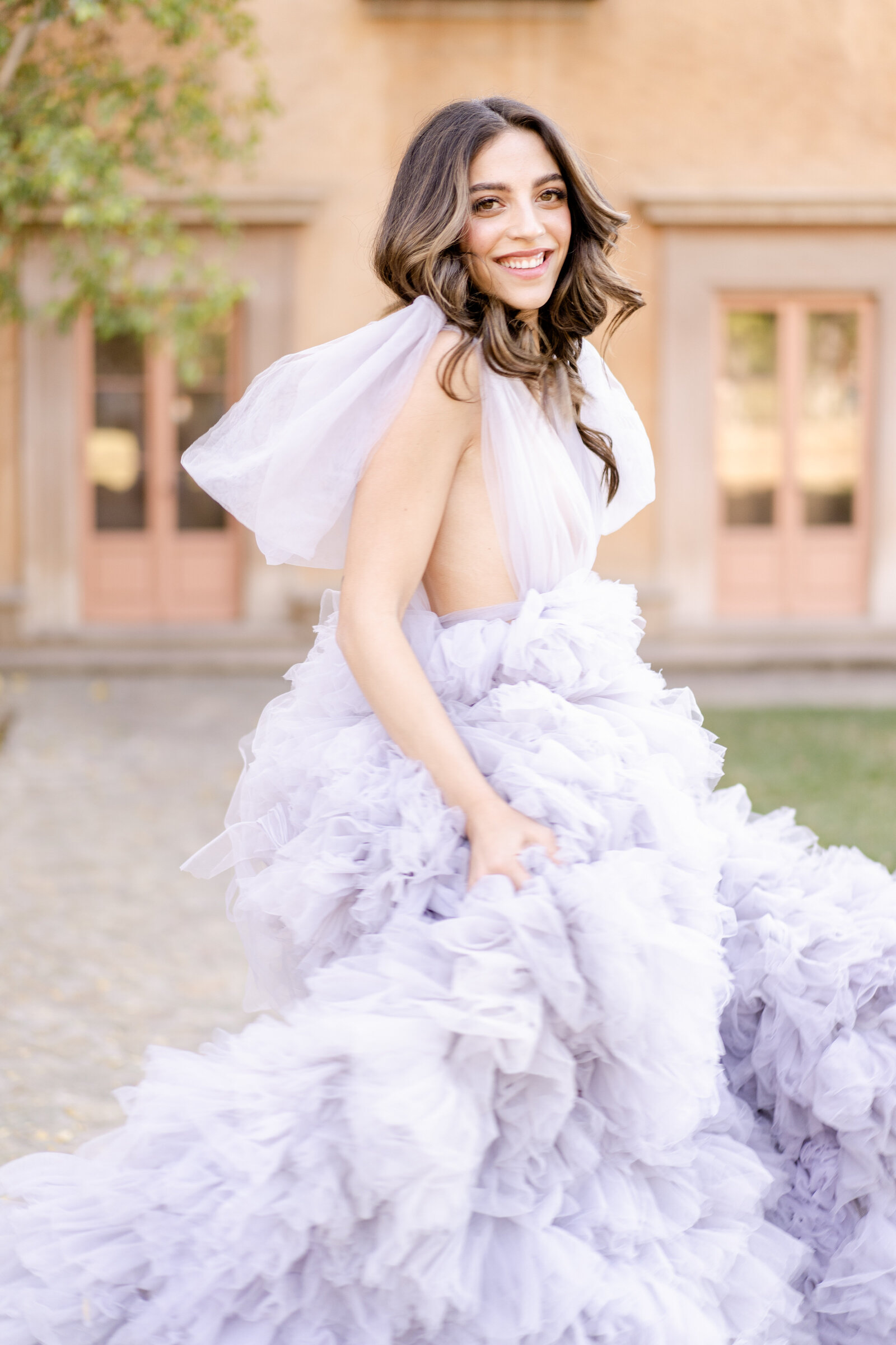 Portrait of a bride in a lavender wedding gown standing in front of a peach-colored building.