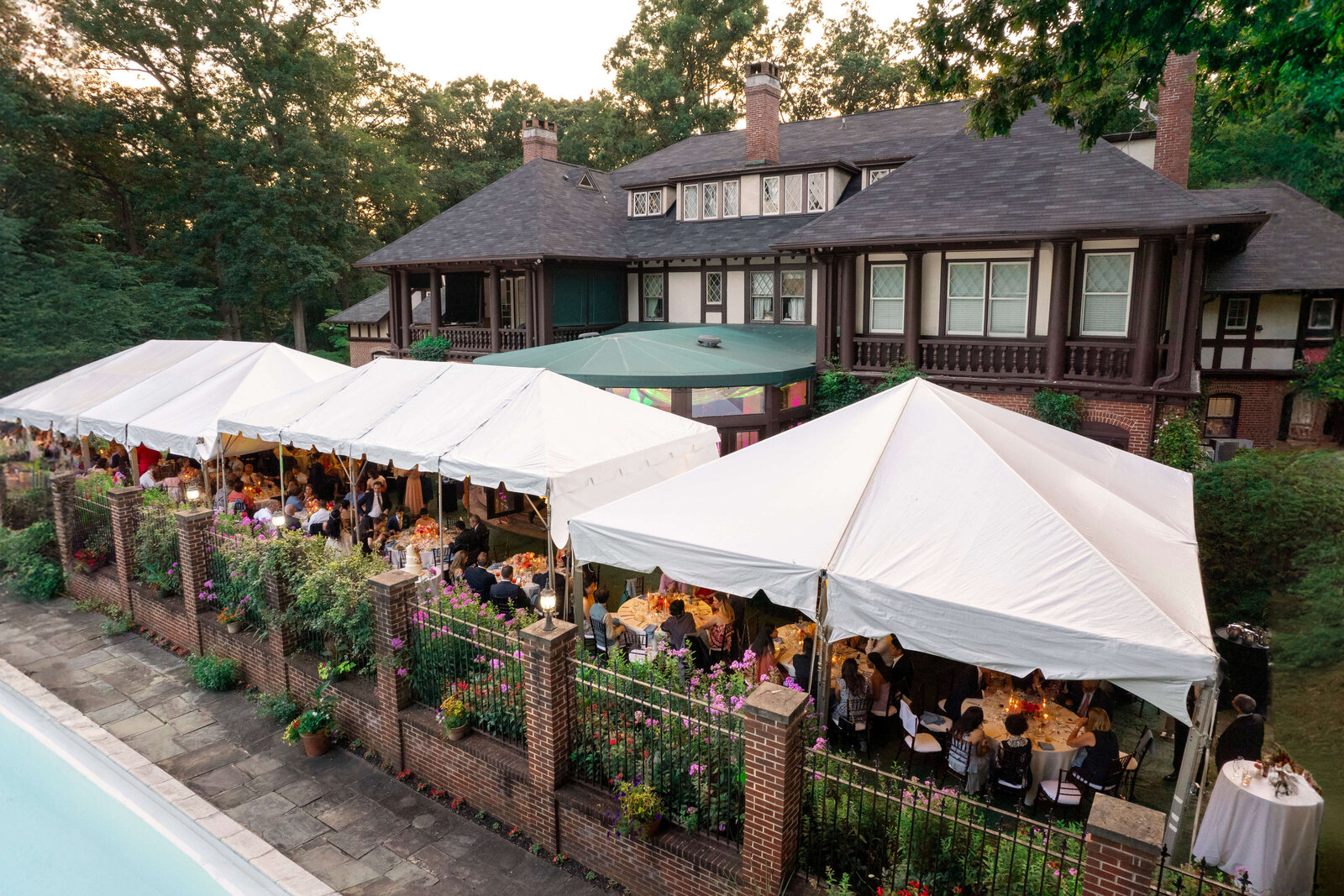 A large outdoor event with white tents is set up beside a Tudor-style house, surrounded by lush greenery. The tents are filled with people seated at tables, and floral decorations are visible along the perimeter.