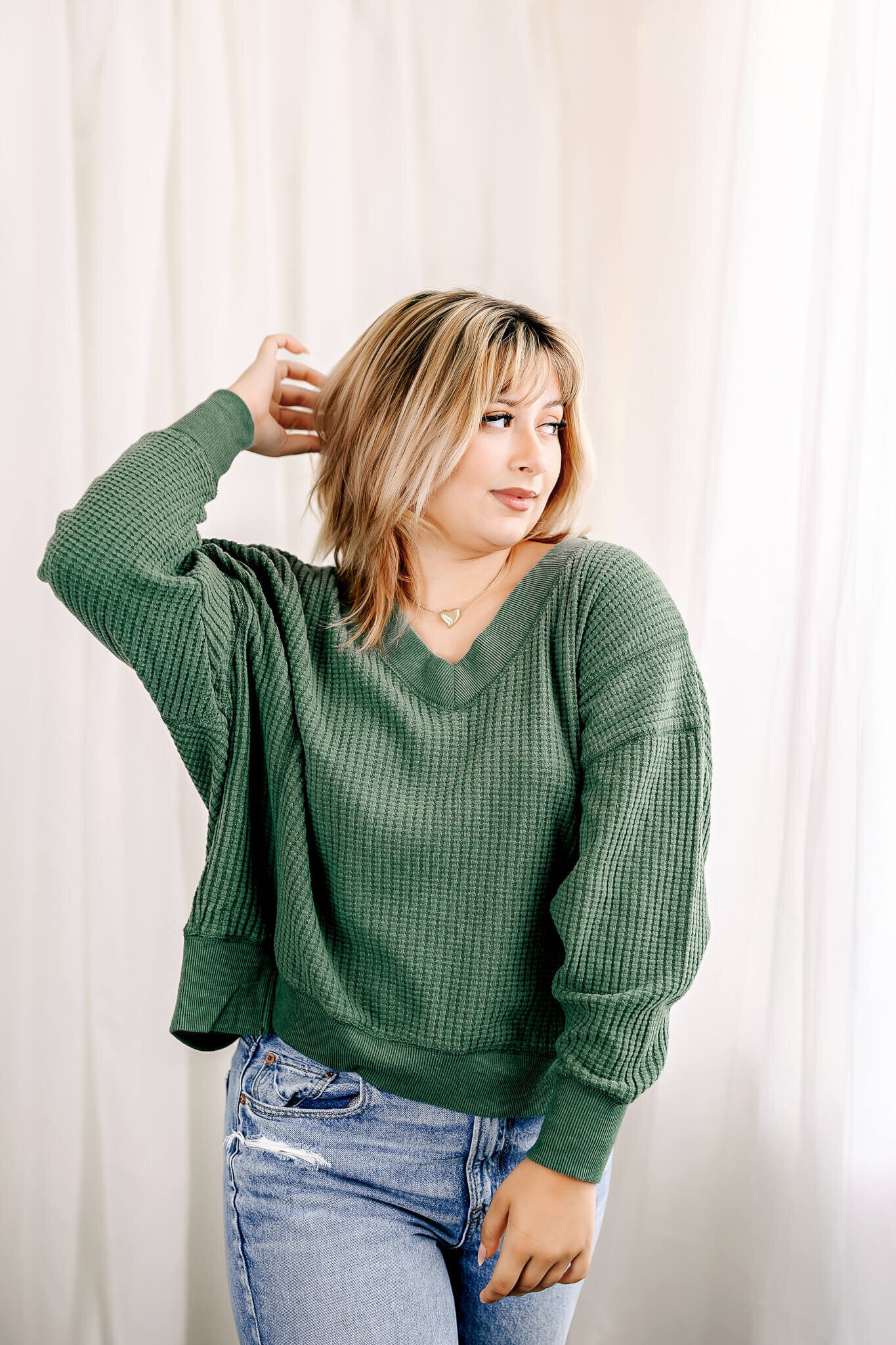 A person with light hair wearing a green sweater and blue jeans poses with one hand behind their head against a white backdrop.