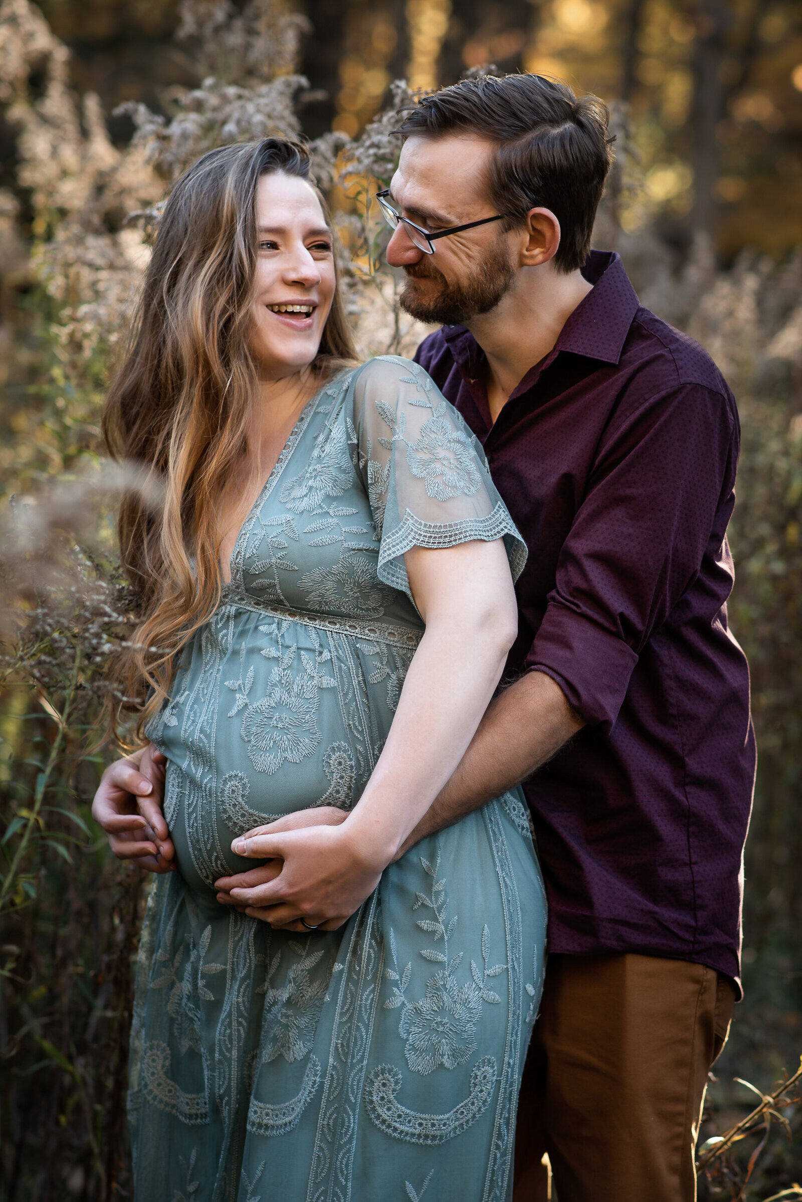 Man holds his wife's pregnant belly from behind while they exchange smiles