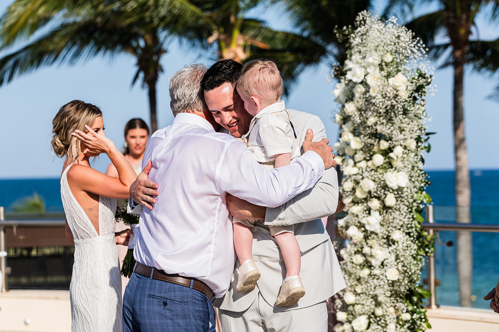 An emotional moment as the groom embraces a family member holding a child during the wedding ceremony, with the ocean in the background. Captured by CMH Studios during a destination wedding