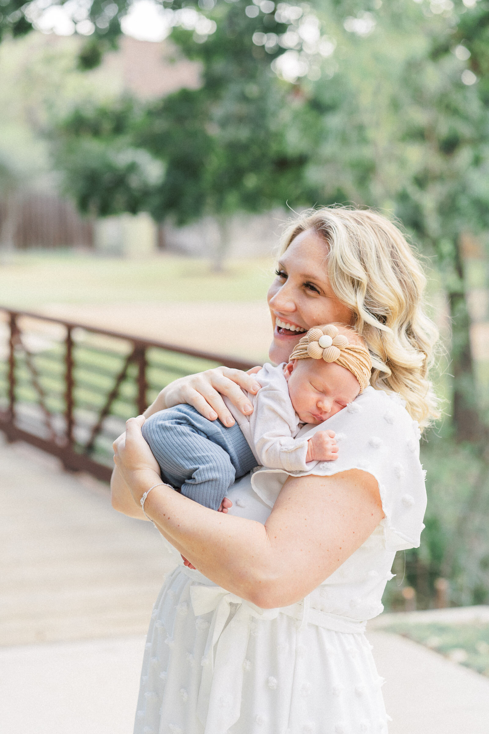 a mother dressed in a white dress stands on a bridge while holding her baby girl. 