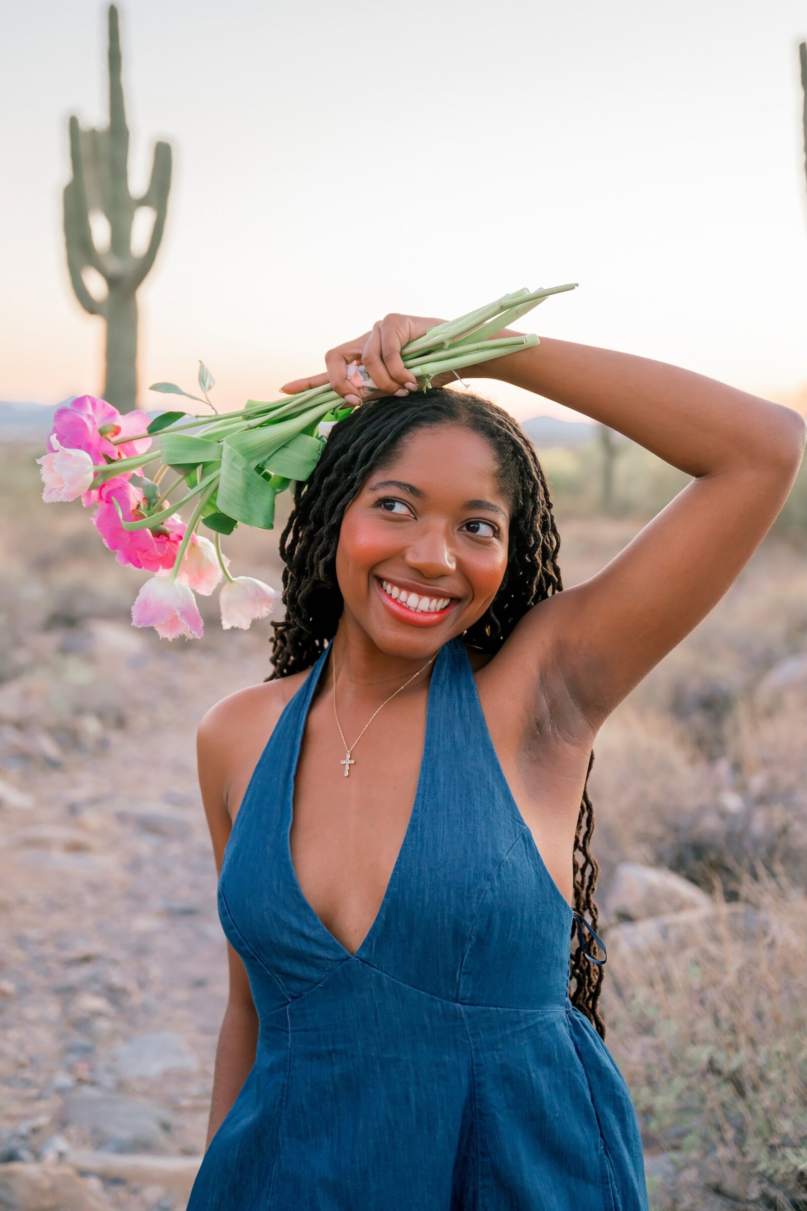 senior girl holding pink flowers over her head in a blue dress smiling looking away from camera 