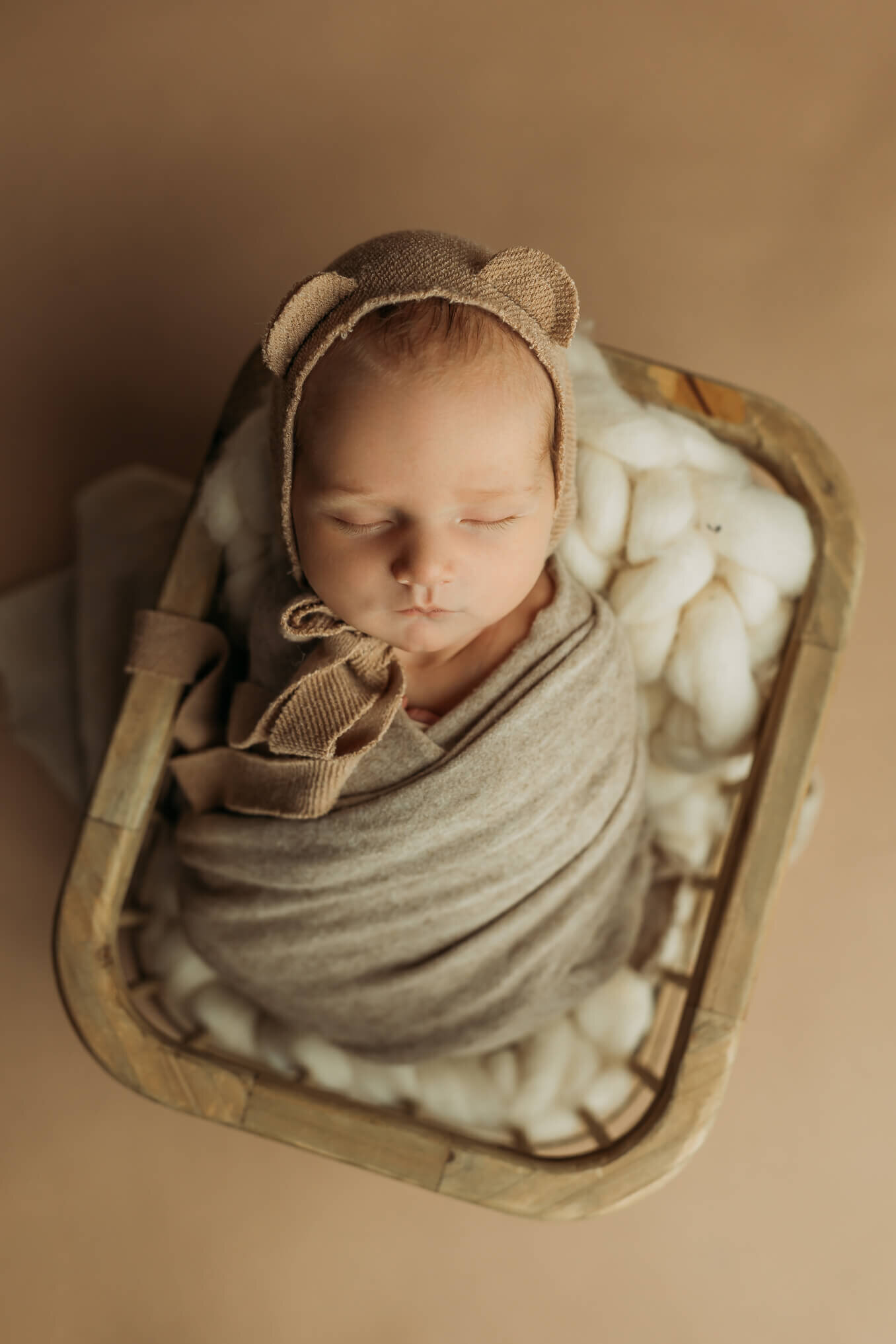 Newborn boy in a bear bonnet wrapped in a swaddle sleeping in a basket on a brown background
