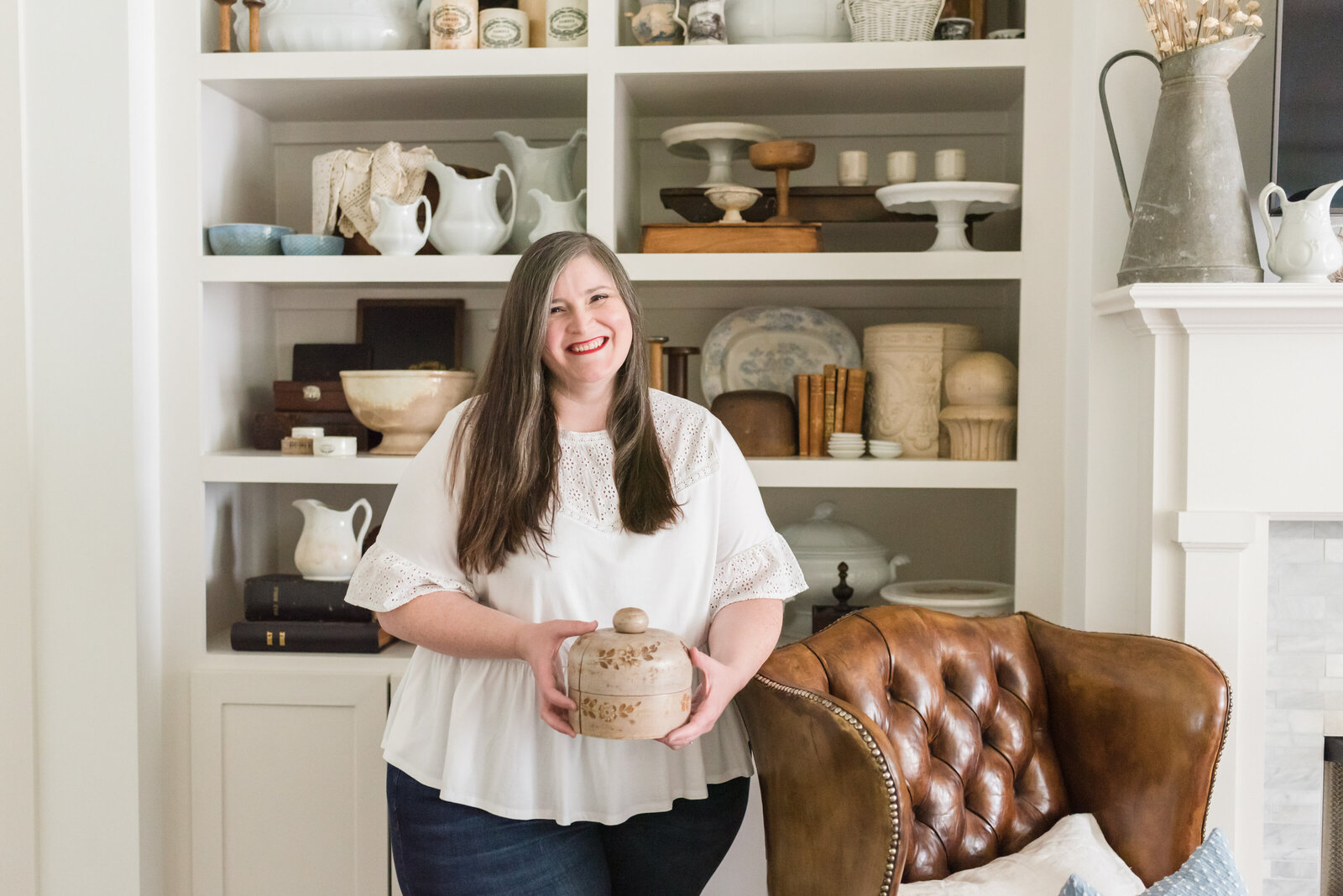 Lauren smiles in front of shelves of her antique collections, resting her arm on leather wingback chair