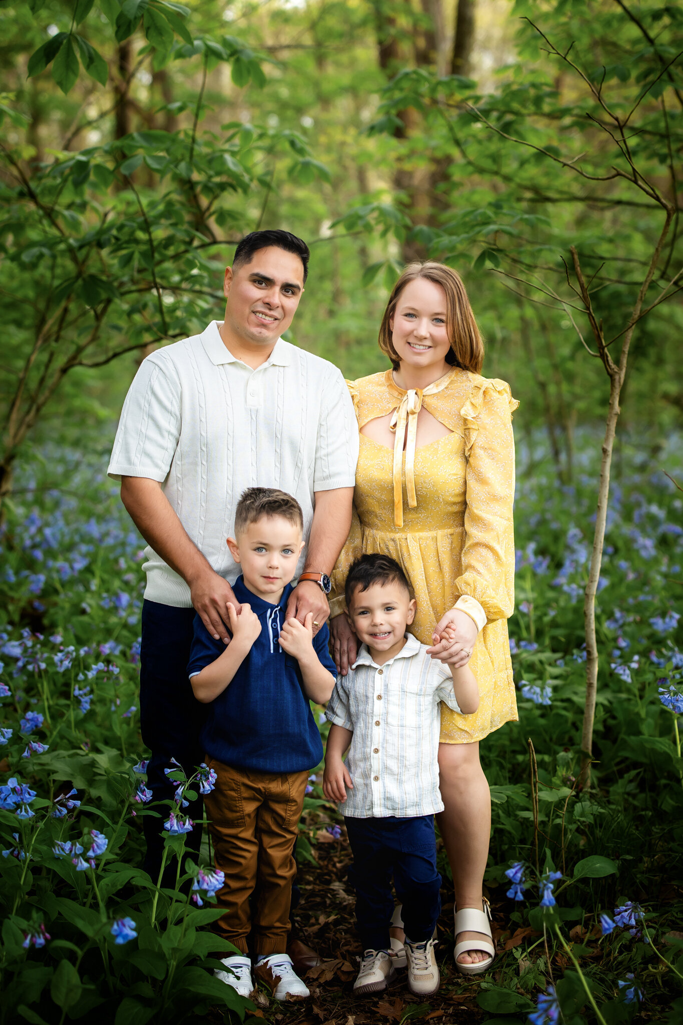 A family of four stands in a wooded area with blue wildflowers, smiling and holding hands. Captured by a Bloomington family photographer, their outdoor portraits showcase joyful moments in nature, dressed in cheerful yellow, white, and blue.