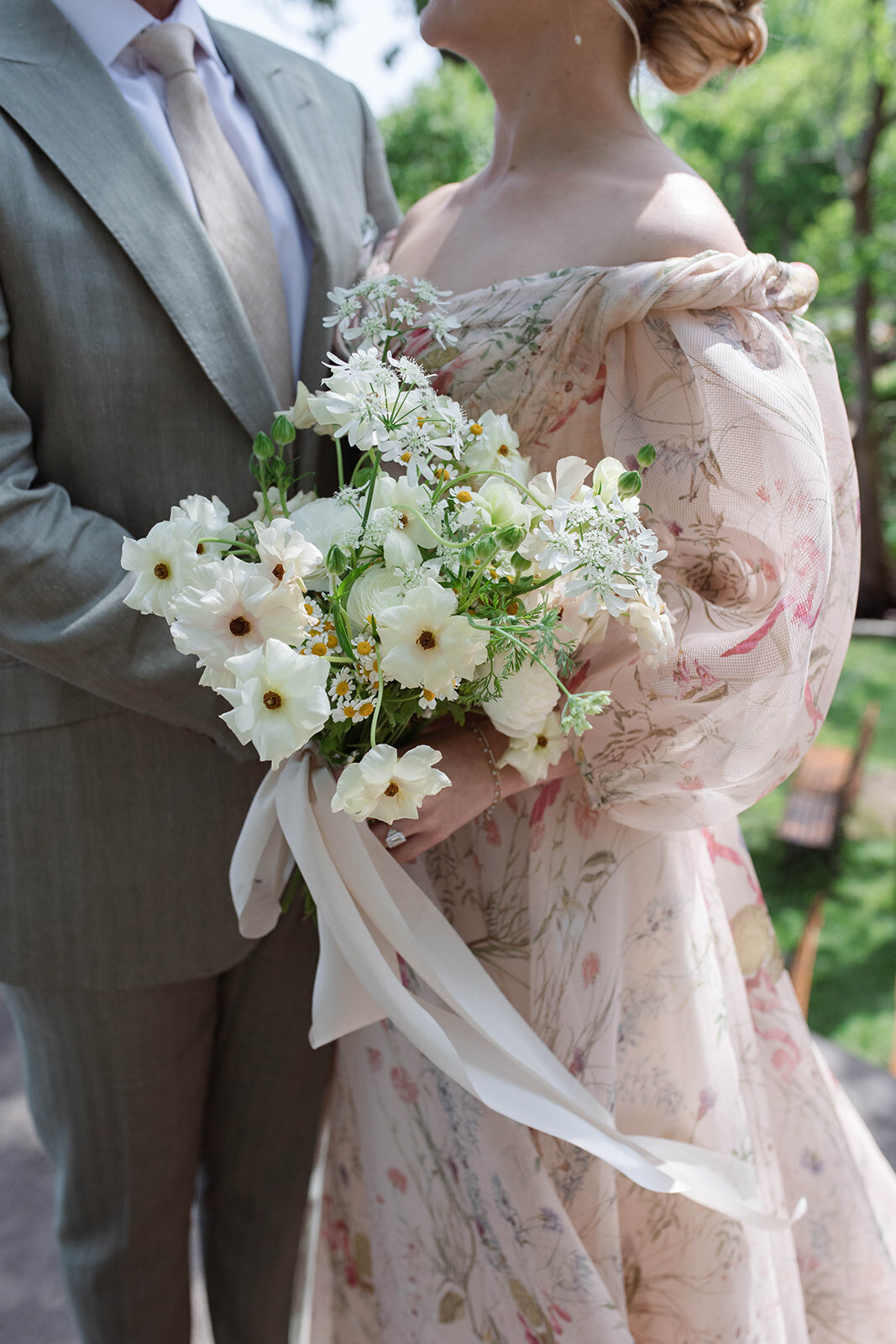 Close-up photo of bride’s floral off-the-shoulder gown. Bride holding garden style bridal bouquet with white and blush butterfly ranunculus and daisies tied blush ribbon.