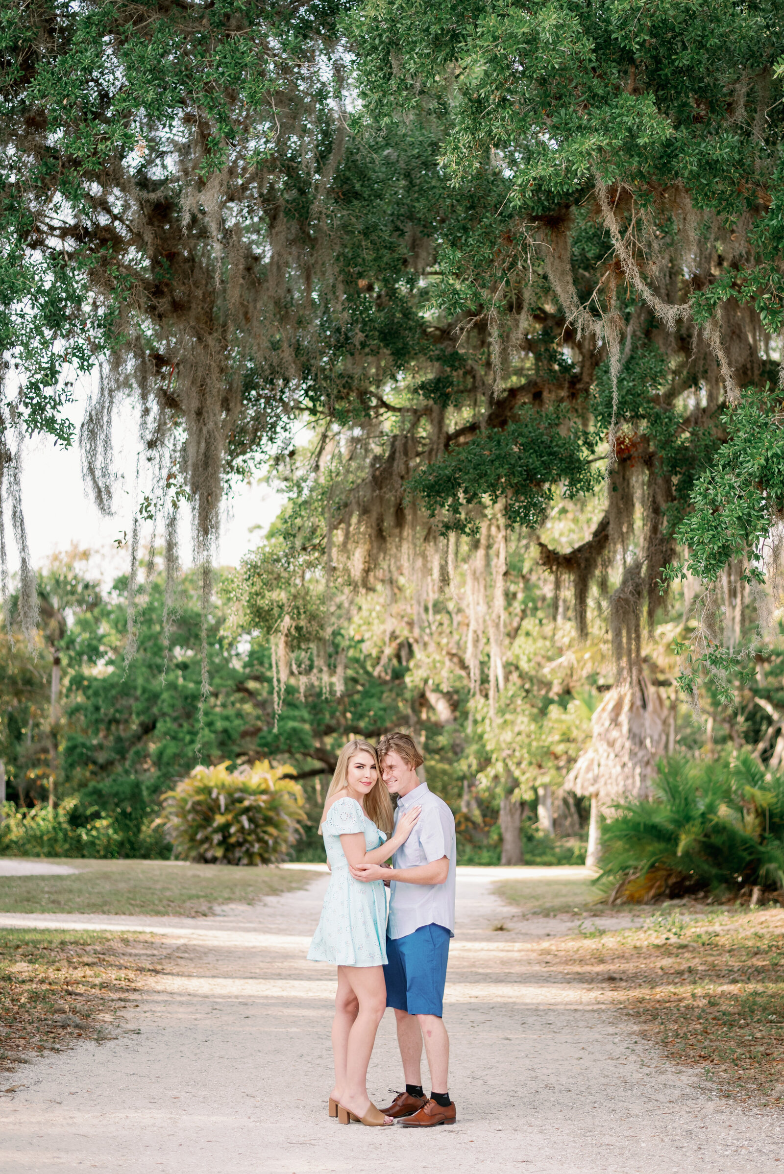 engaged couple standing front to front, arms and hands on each other. Woman is looking at the camera smiling while her finacé looks slightly at her. They are on a sandy pathway with a large mossy oak tree above them