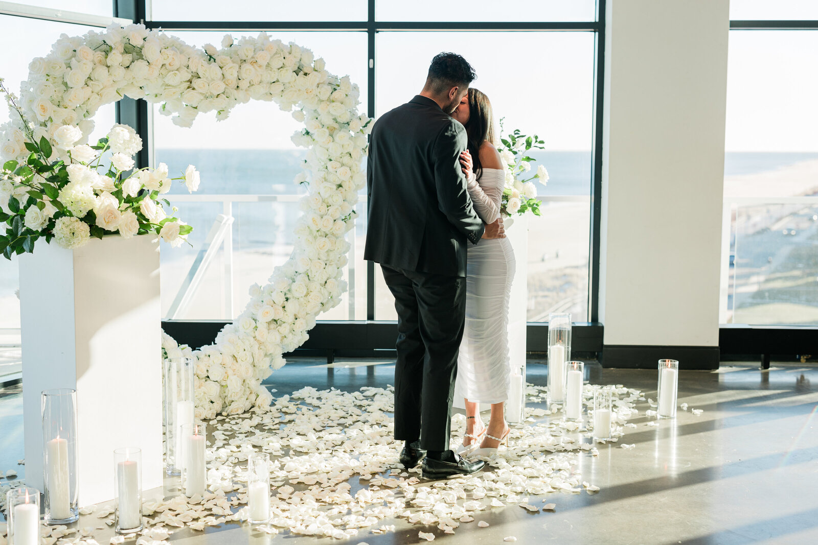 Couple kissing in front of a white floral heart arch with petals and candles, overlooking the ocean, styled by Yours Truly, a wedding planner and designer in New Jersey.