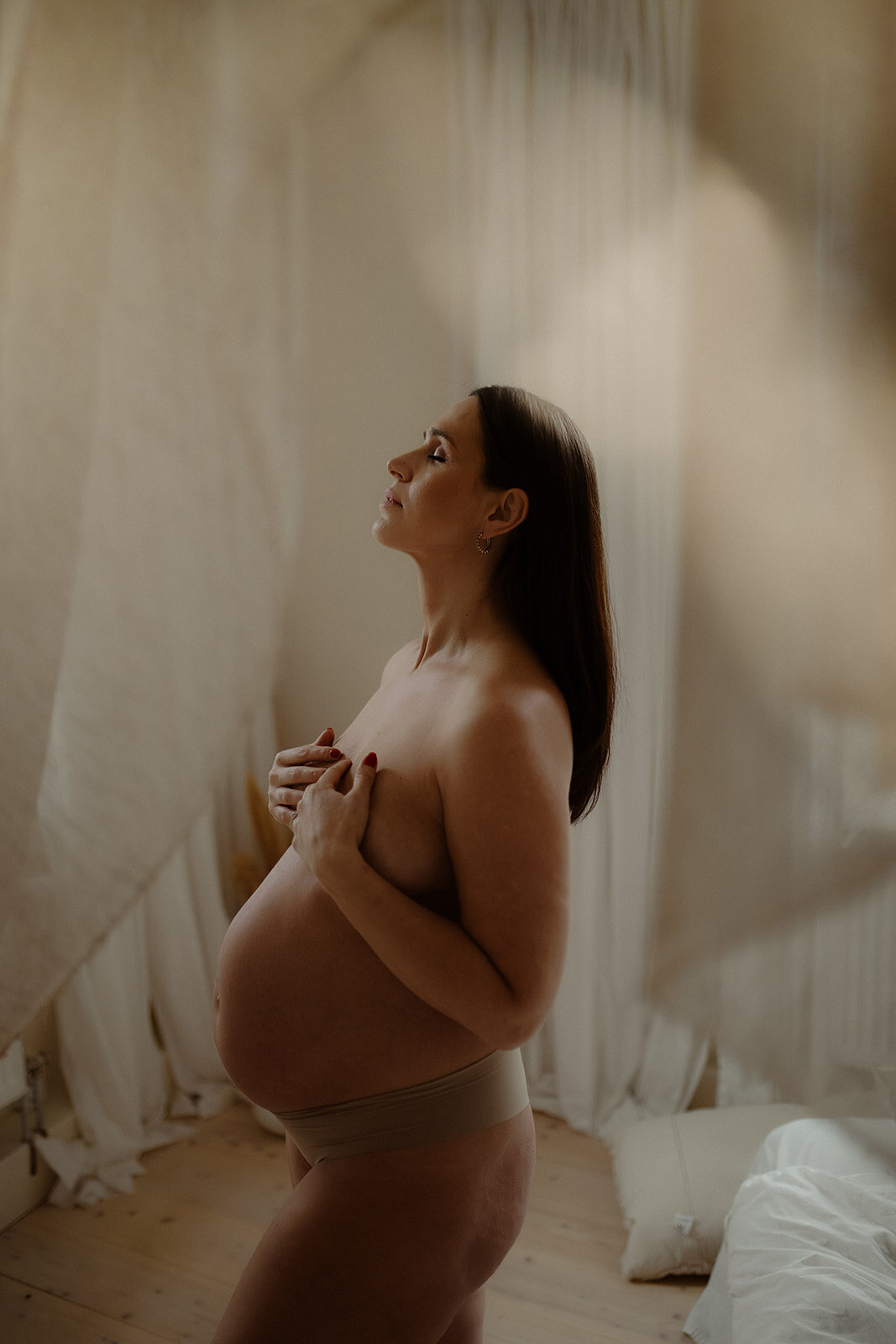 en gravid mamma står i trosor i ett sovrum och håller om hennes bröst
