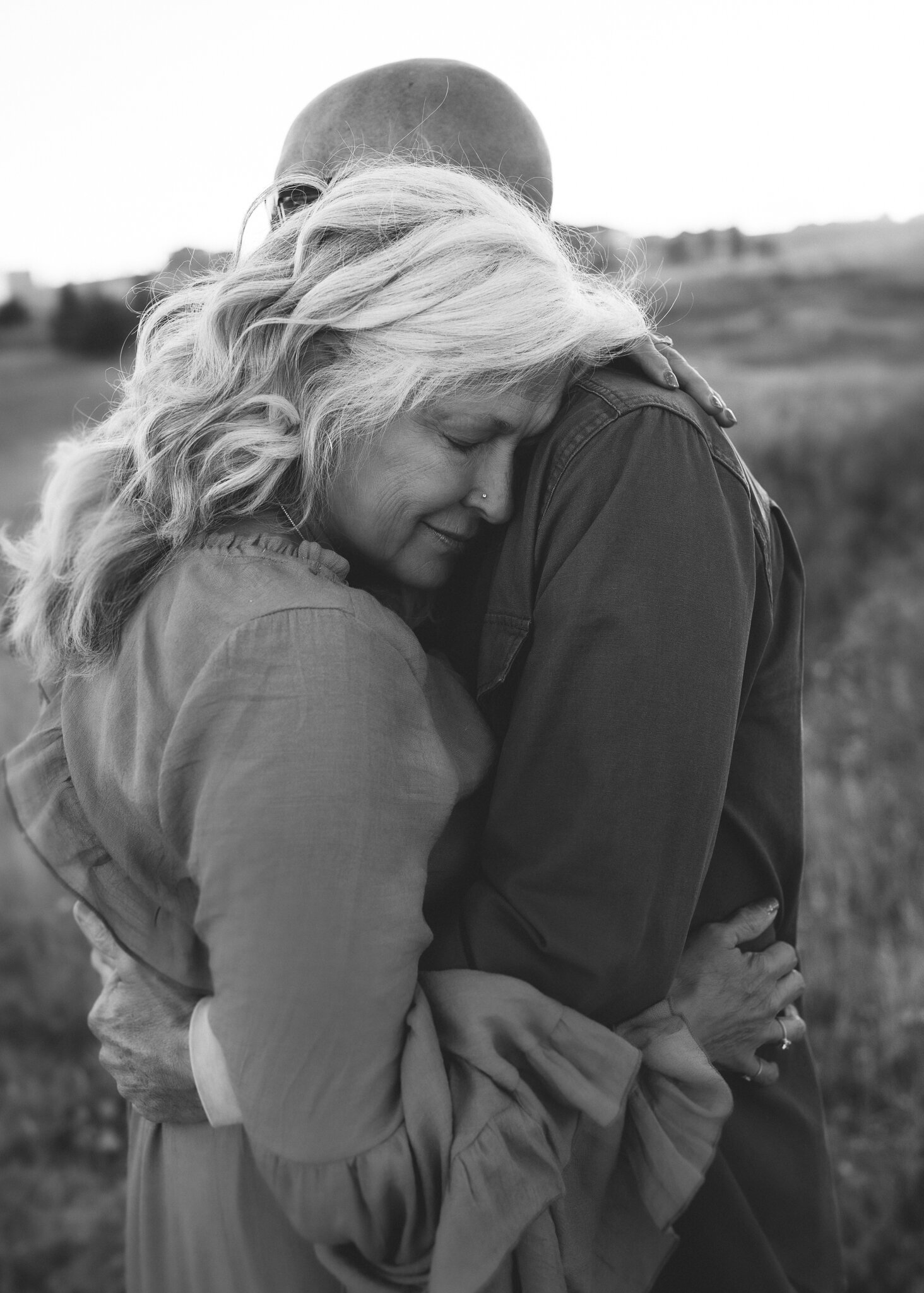 A senior couple embraces during an outdoor couples session. The woman has striking white curly hair and she lays on her husband's chest as he holds her close.