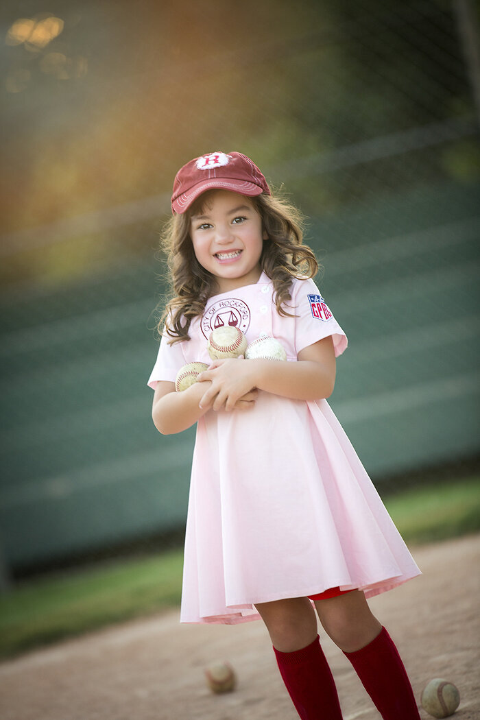 Girl holding baseballs in her arms in baseball field, a Dallas child photographer.