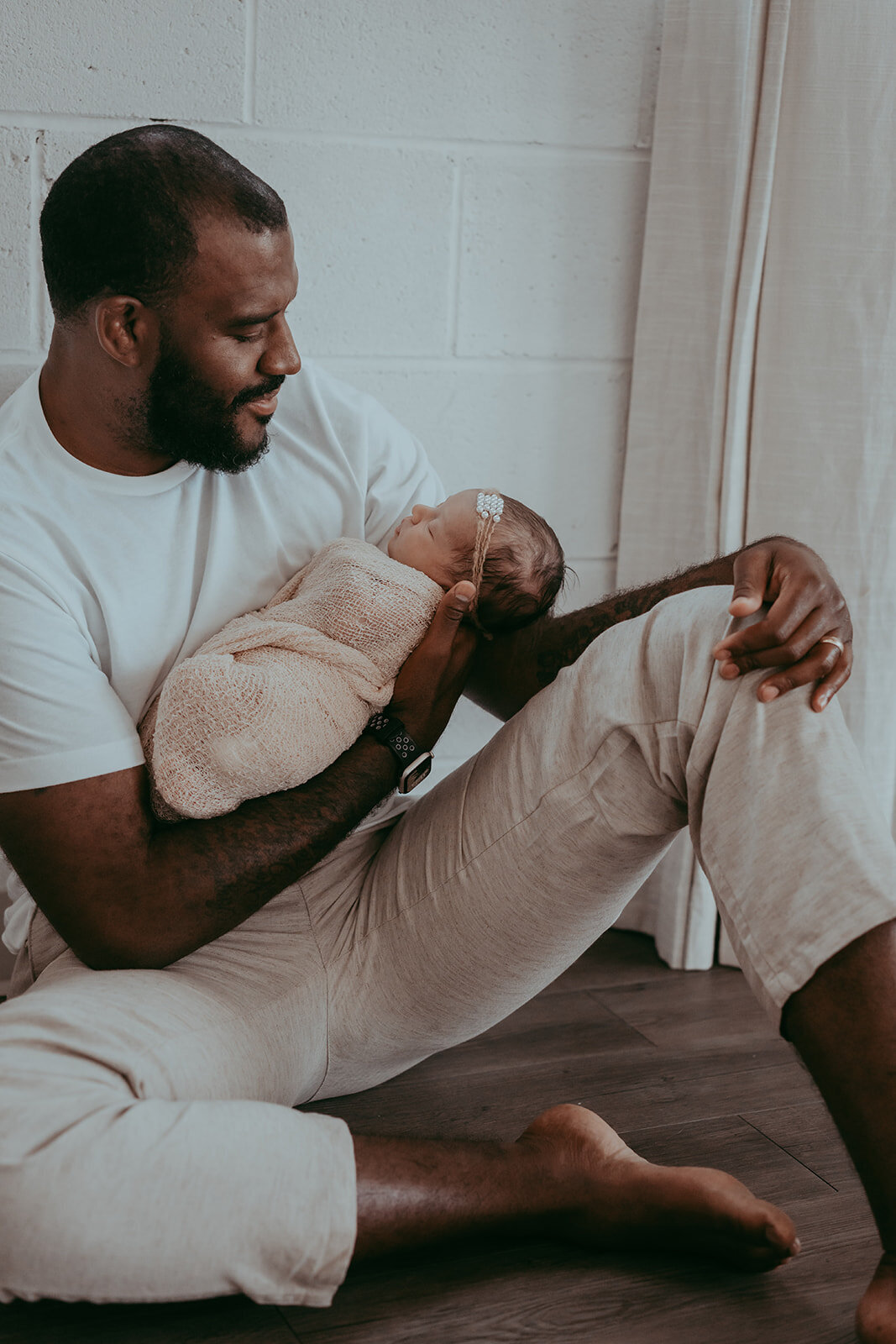 father leans agains exposed brick wall while holding newborn in one arm