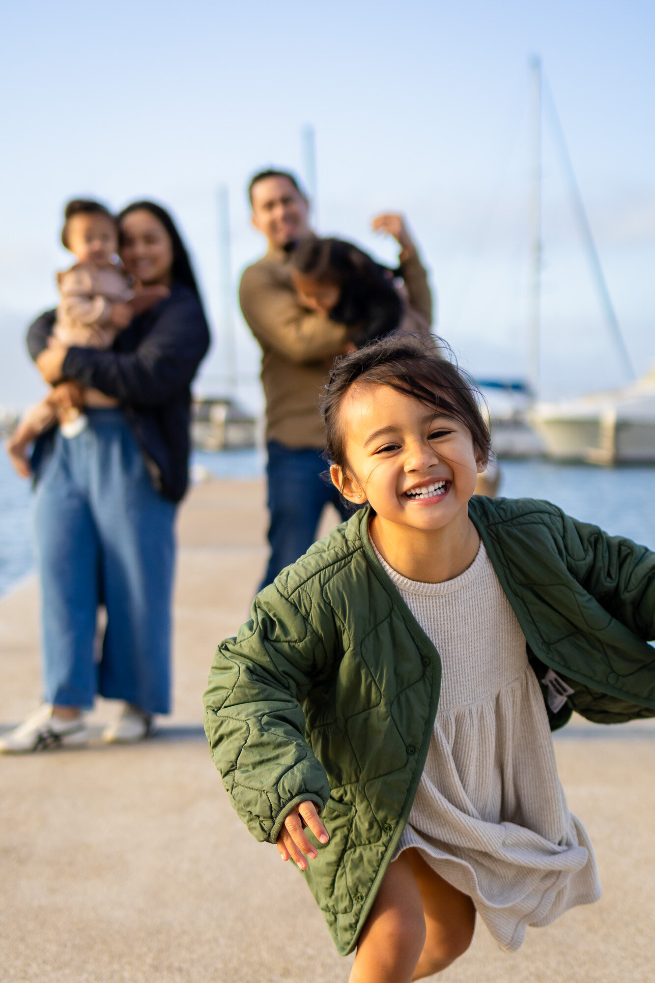 Joyful young girl runs toward the camera with her smiling family in the background during a candid dockside session — Ellobelle Photography Family Photographer.