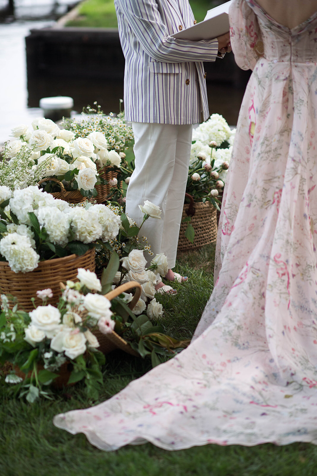 Wicker baskets filled with white garden roses, white hydrangea, blush peonies, and blush butterfly ranunculus. Close-up of the bride’s floral gown and an officiant wearing a pinstripe jacket.