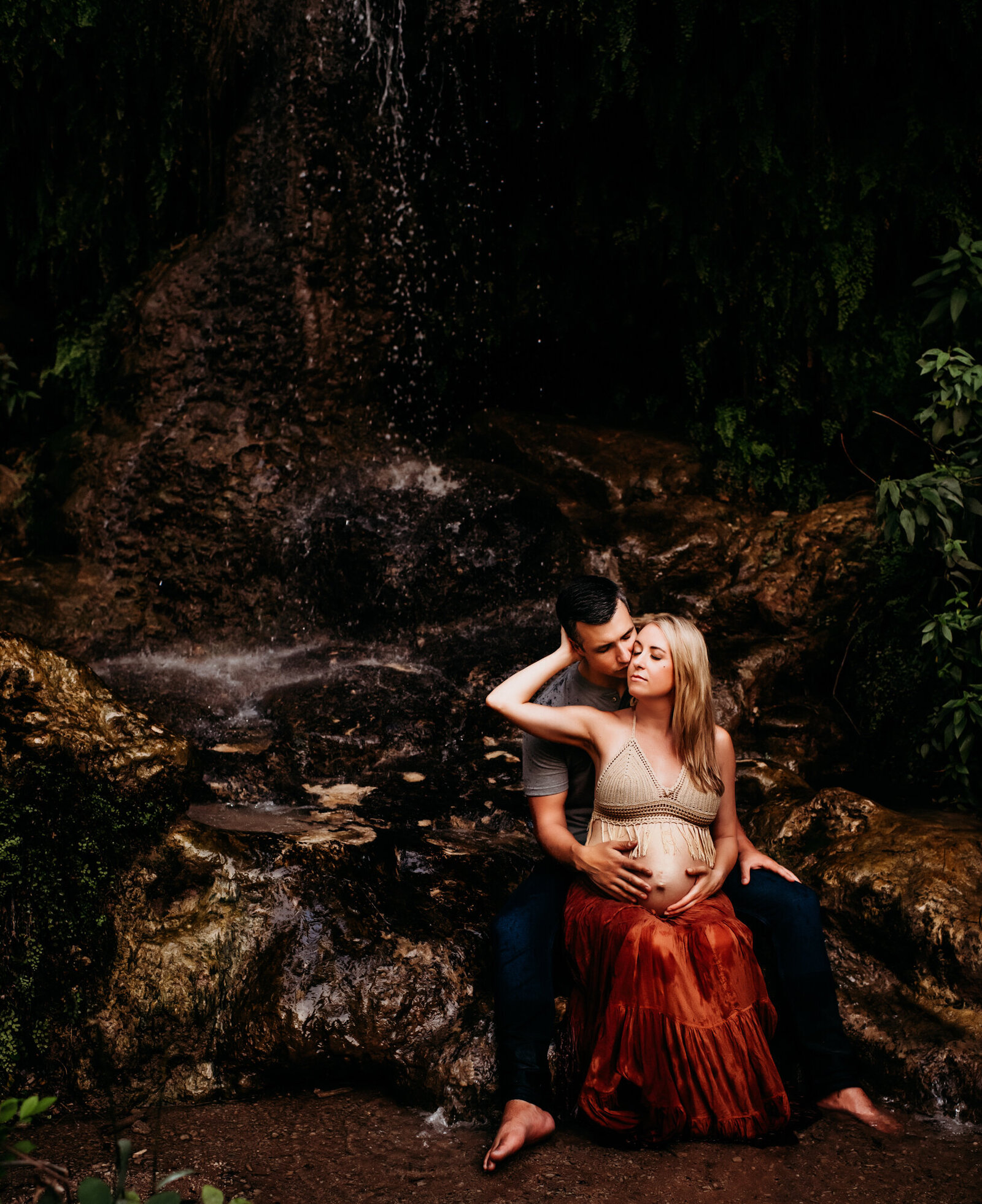 Maternity Photographer, expecting mother sits in husband's lap near a waterfall in the forest