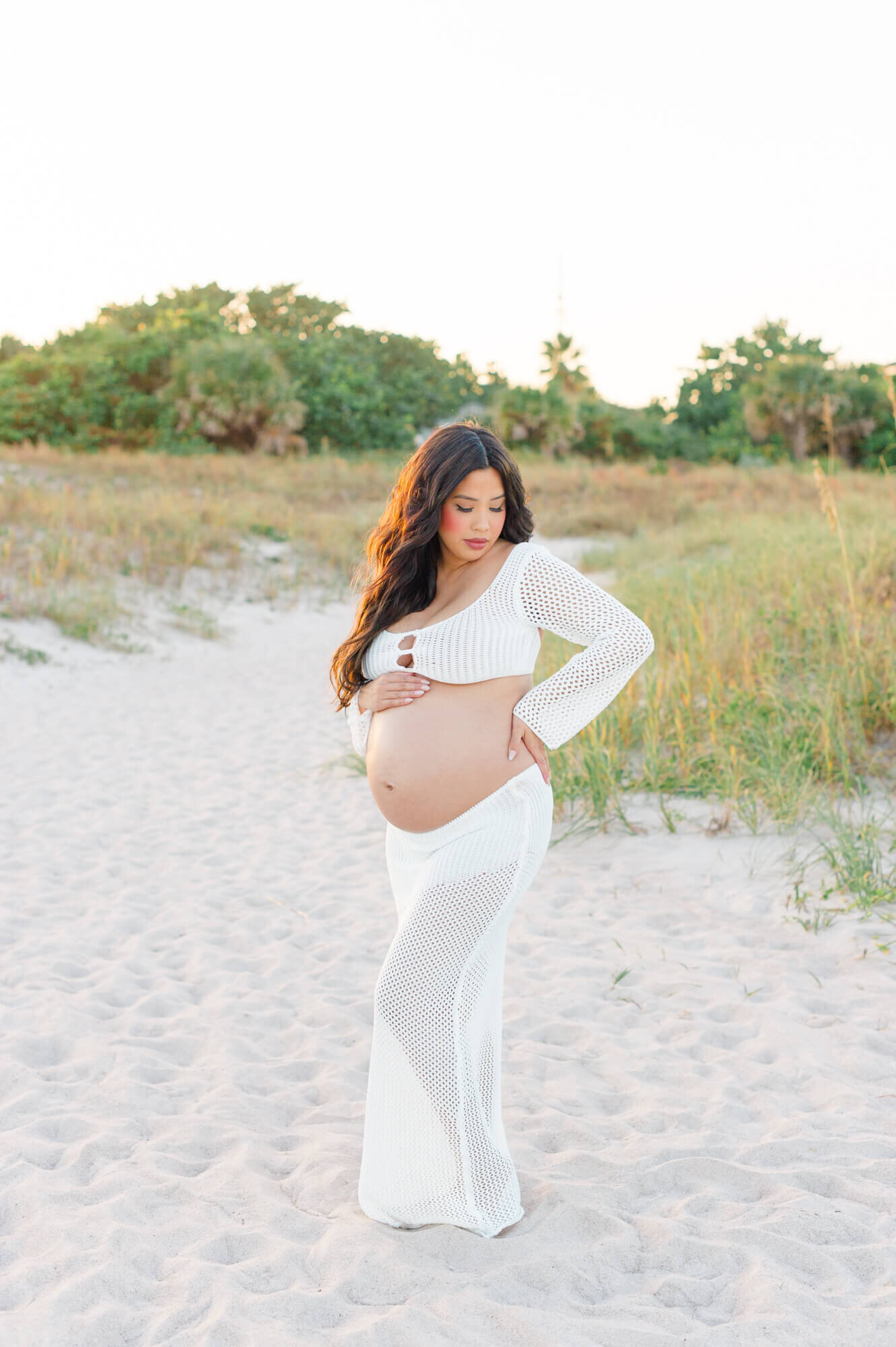 Pregnant mom stands in knit white two piece while holding her belly and looking down during her Orlando maternity photo session