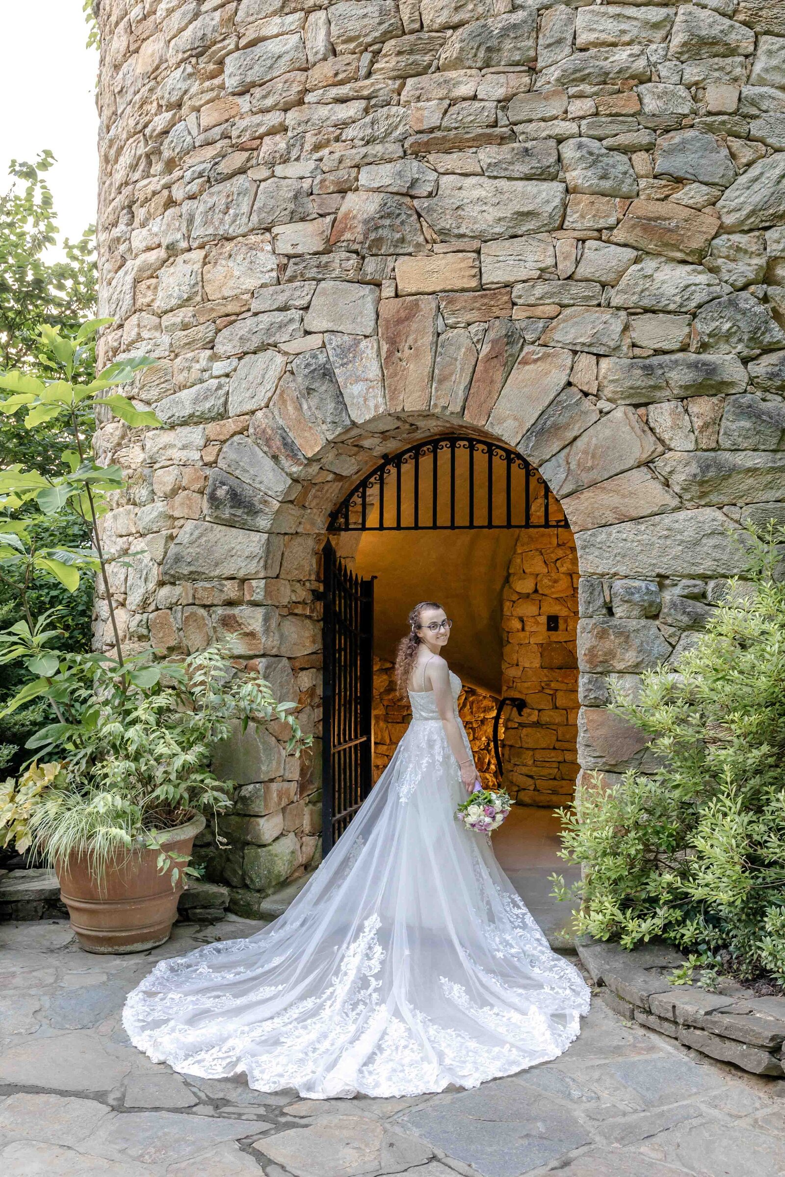 Bride in a breathtaking lace gown standing gracefully at the entrance of a rustic stone tower in Longwood Gardens, highlighting elegance in ennett Square, Pennsylvania.