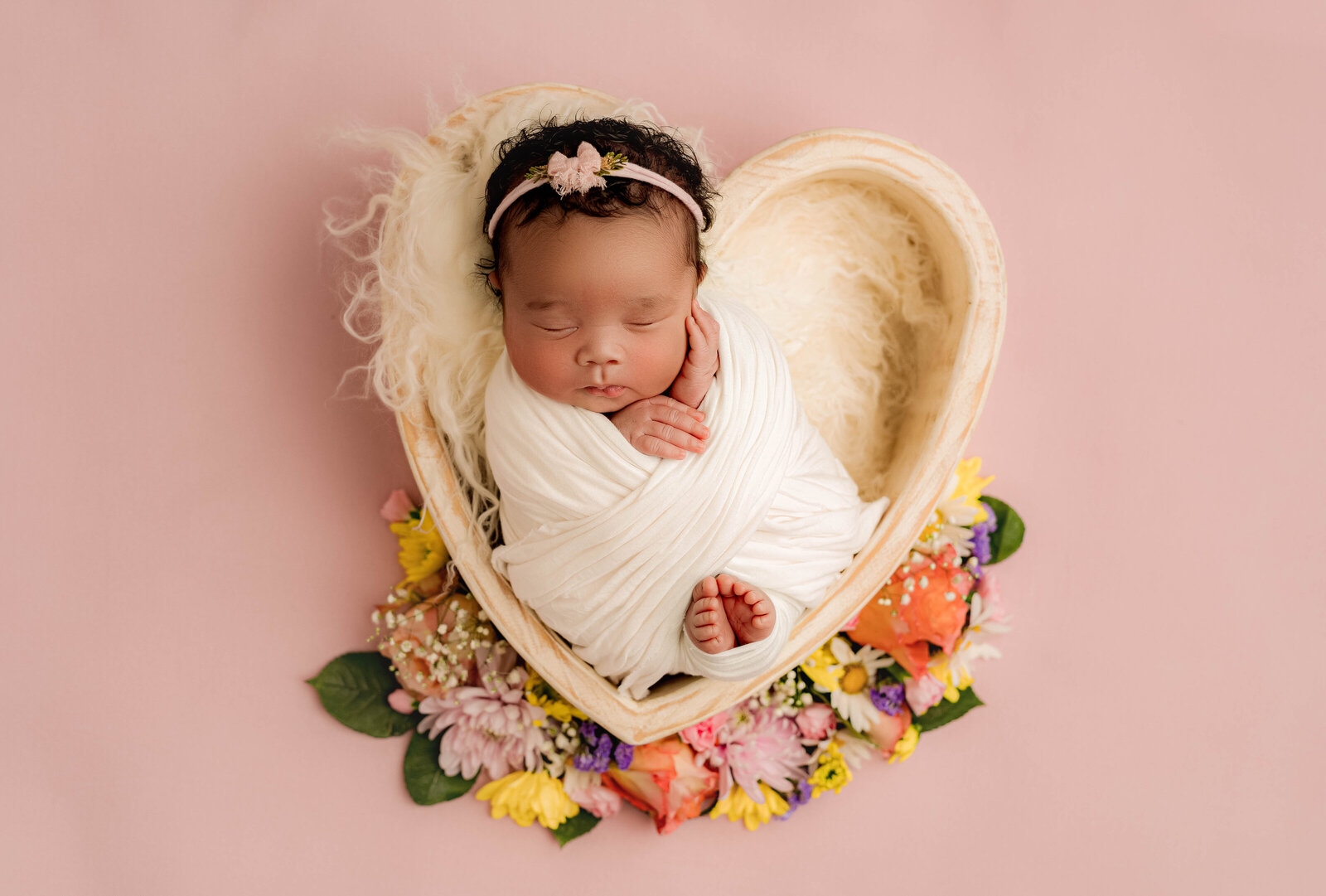In a serene Charlotte newborn photography studio, a baby sleeps soundly wrapped in a white blanket on a heart-shaped bed lined with soft fur. The pink backdrop and colorful flowers add charm, while the floral headband completes the peaceful scene.