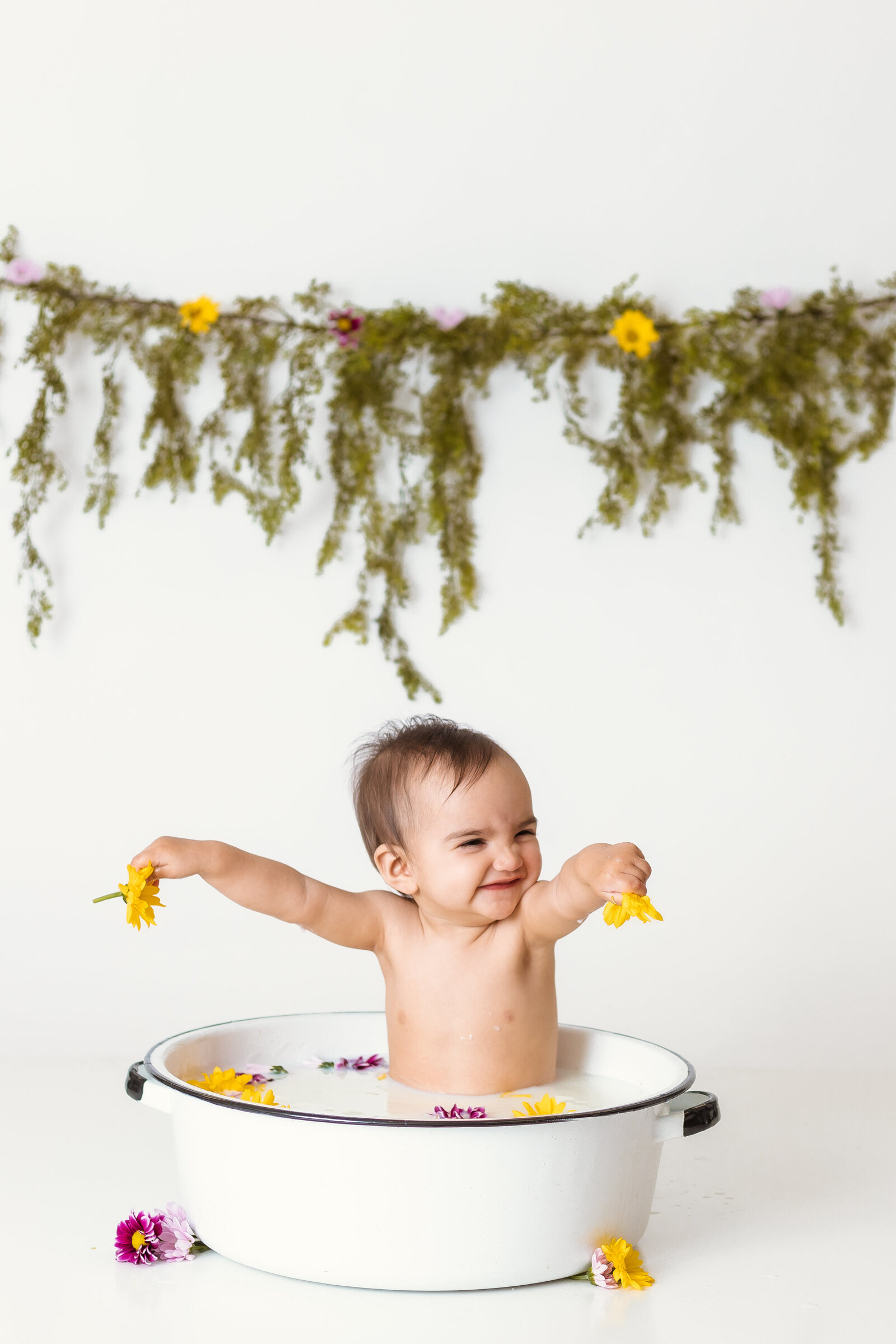 Milestone Photographer, a baby sits in a ceramic pot holding flowers in a milk bath