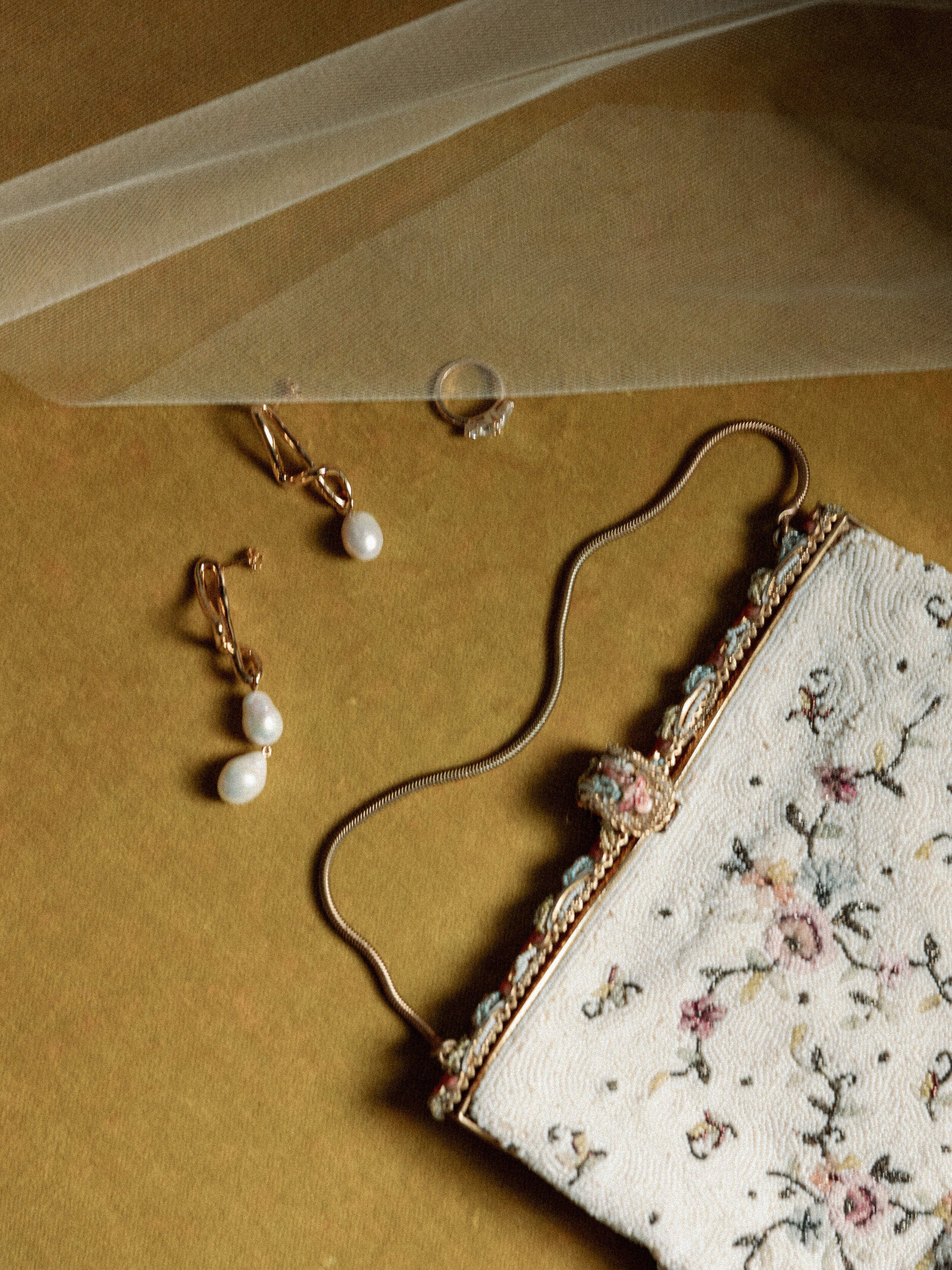 Retro style bridal accessories with pearl earrings, beaded floral clutch, and veil on top of mustard colored velvet couch and dramatic window light 