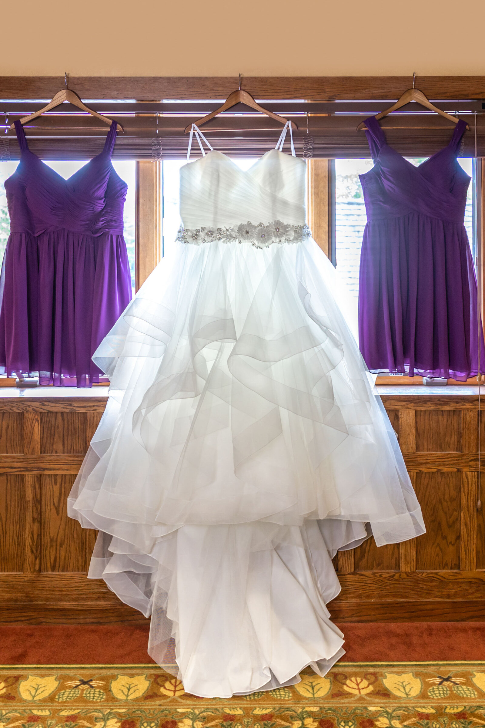 A white layered wedding dress with sparkling belt detail is displayed between two purple bridesmaid dresses in front of a wooden window, captured during a Boone, NC wedding, showcasing elegant bridal attire.