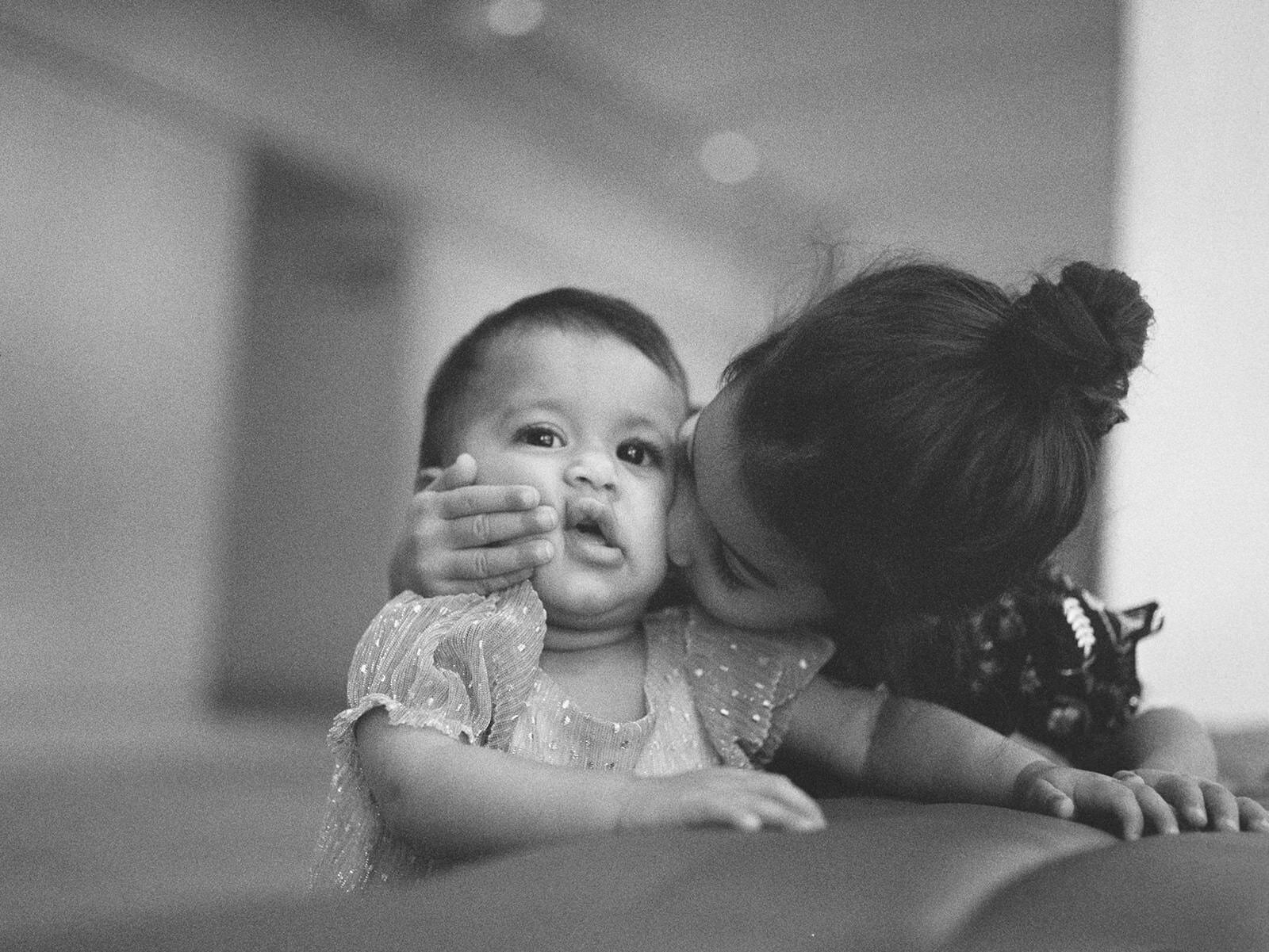 A tender black-and-white moment of a young girl lovingly kissing her baby sibling on the cheek, both dressed in elegant outfits."