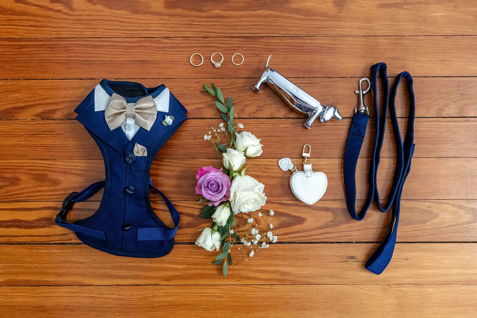 A creative flat lay of wedding day dog accessories, including a navy tuxedo harness, leash, floral boutonniere, and wedding rings, perfect for a pet-inclusive wedding in Coatesville, PA.