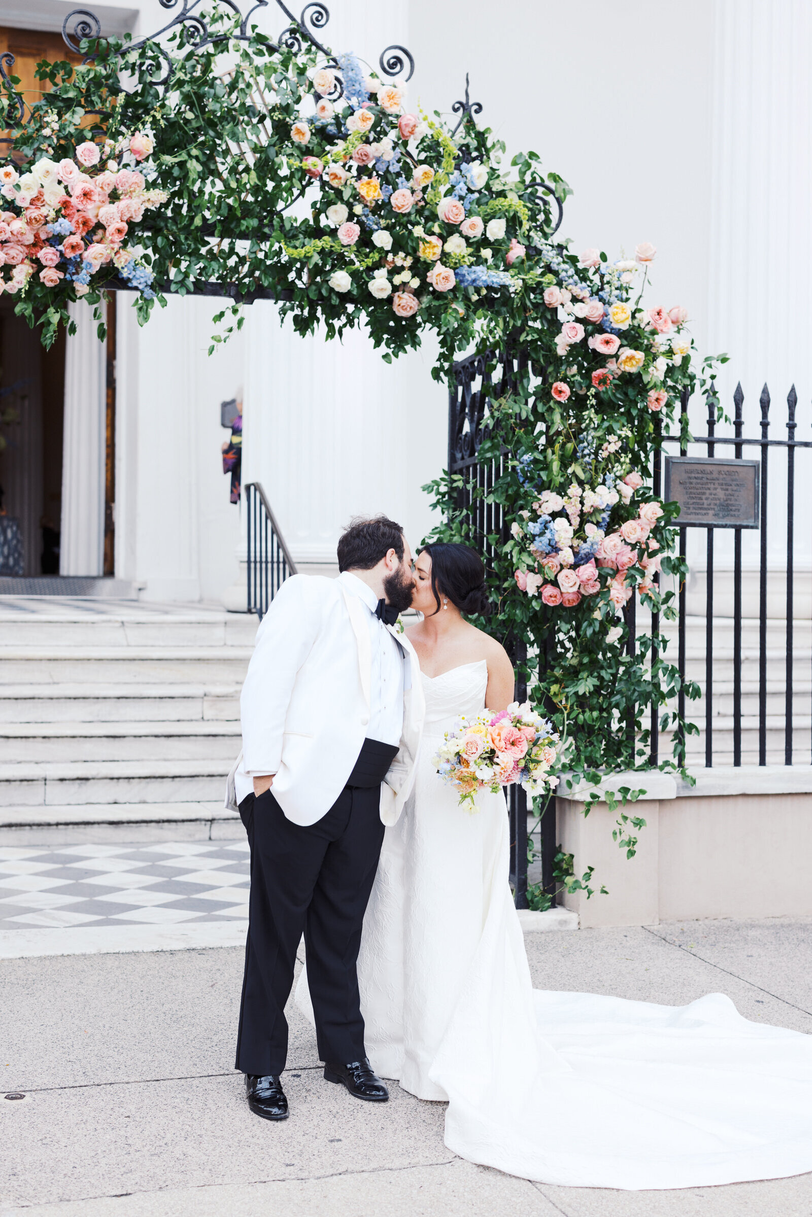 A bride with a pink wedding bouquet and a groom in a white tux kiss in front of a colorful floral arch at Hibernian Hall in Charleston South Carolina