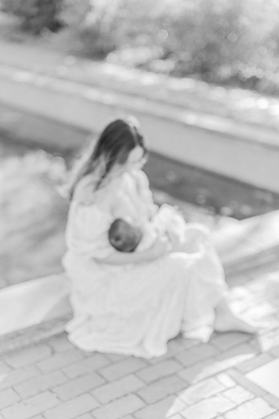 A black and white image with a subtle intentional blur of a mother  breastfeeding her baby girl while seated outside at a reflection pool at the Arlington Town Hall