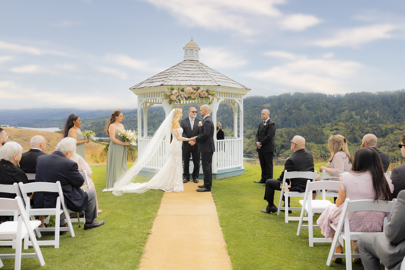 Bride and Groom exchange vows during  a wedding ceremony with guests looking on in front of green mountains and a gazebo. Wedding photographer philippe studio pro from Sacramento captured the moment with his camera.