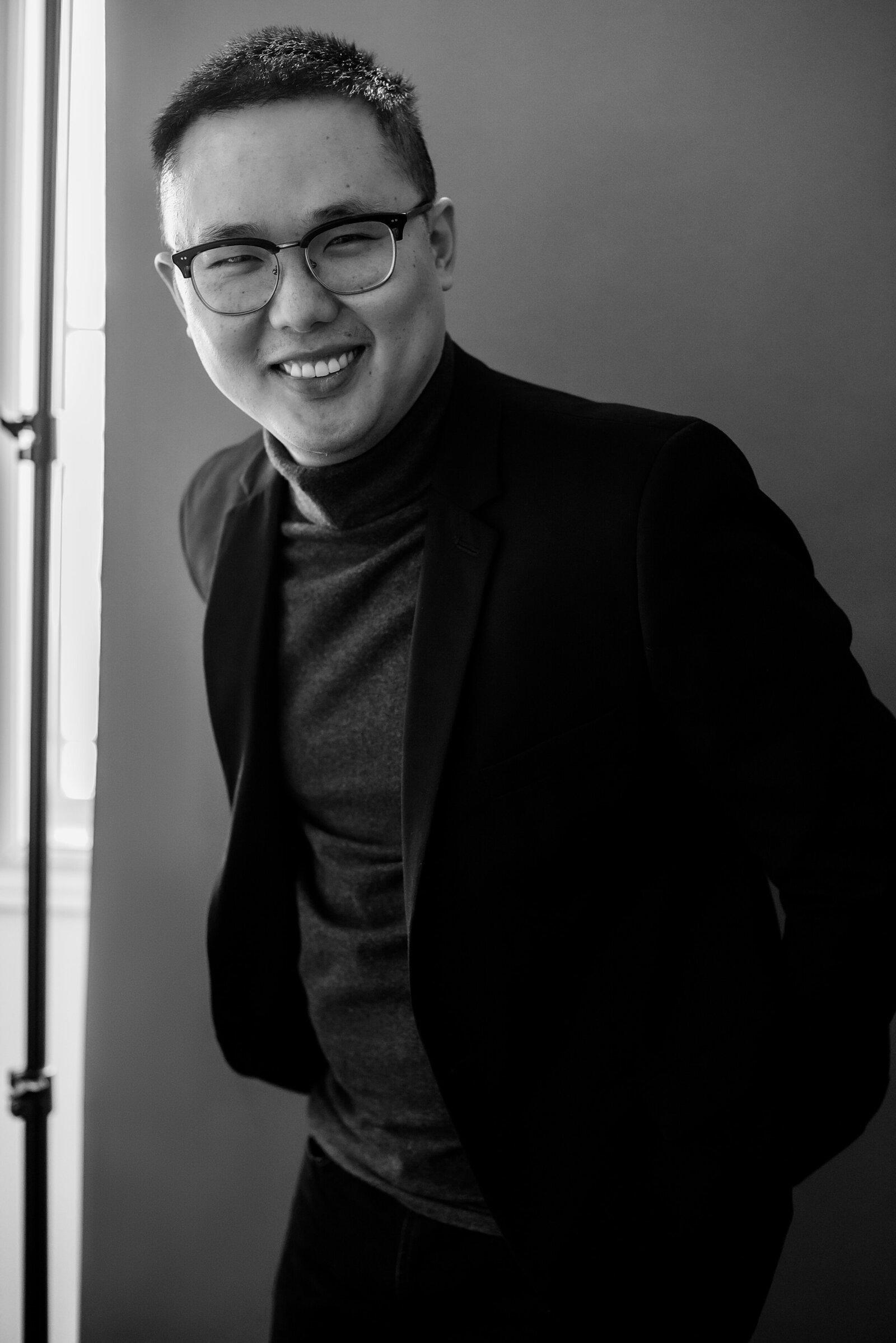 Black and white editorial headshot of an Asian man in  a dark suit and glasses