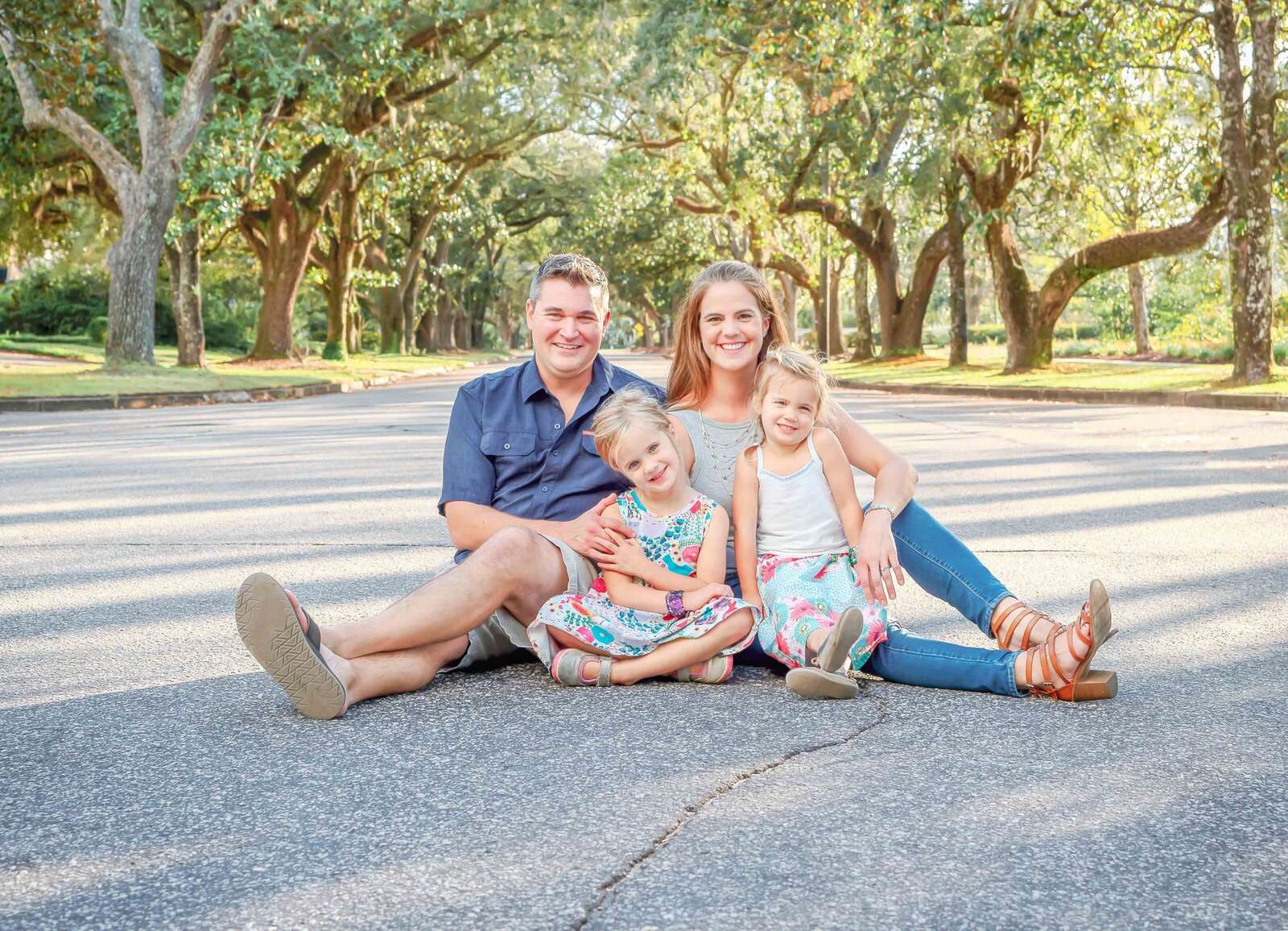 A family of four sits together on a tree-lined road, dressed in casual summer outfits. The dappled sunlight and lush trees create a warm, inviting setting, perfect for destination family photographer NC, highlighting joyful and candid family interactions.