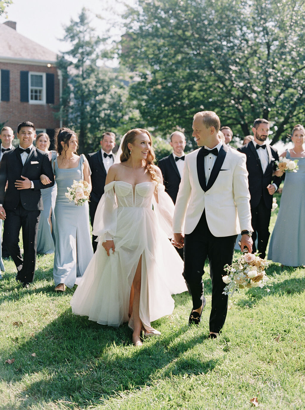 Bride and groom walking in front of the bridal party outside of the Brittland Estate mansion wearing a white tuxedo and an off the shoulder dress.