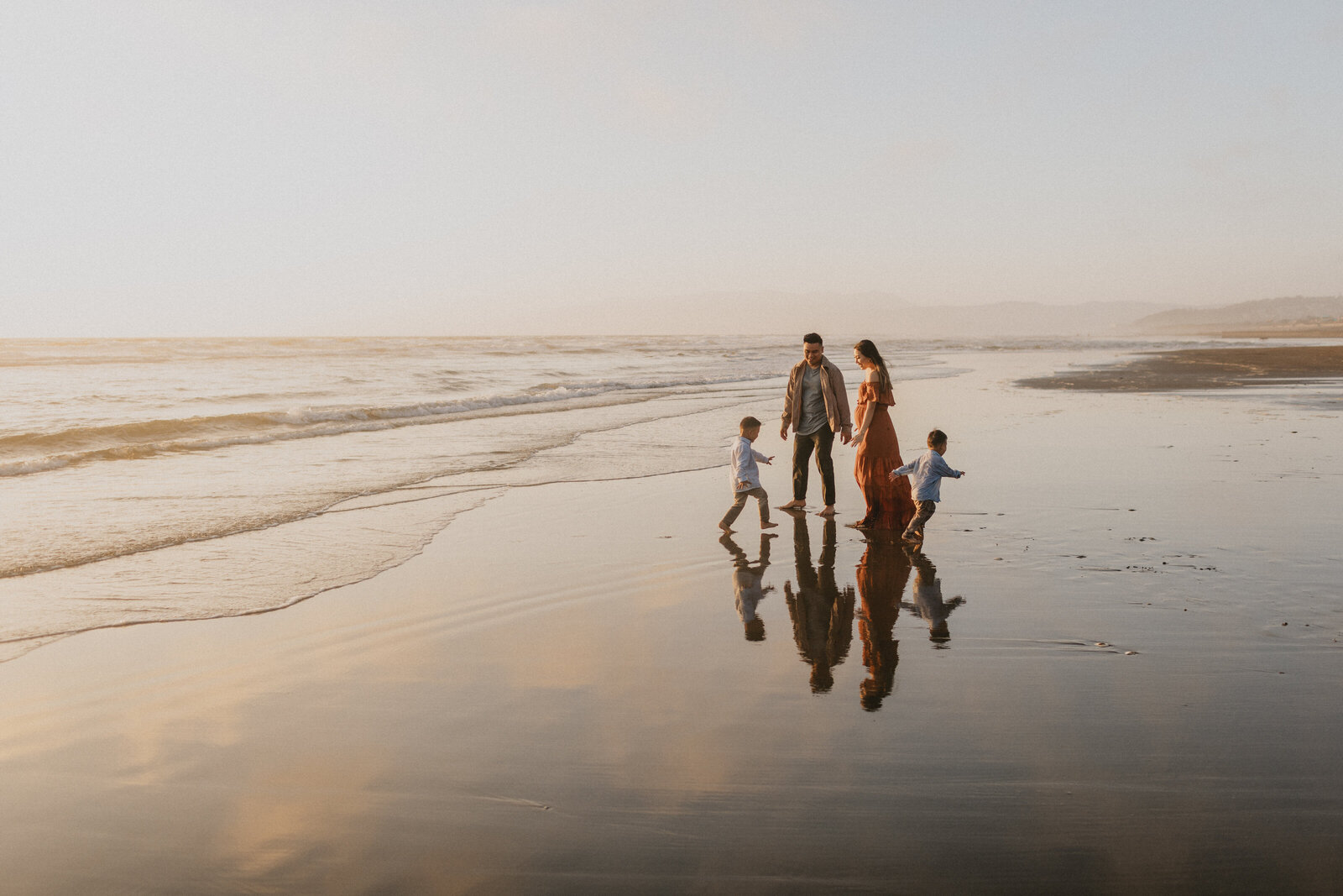 A family of four walks along a beach at sunset, their reflections visible on the wet sand as gentle waves roll in. two adults and two children enjoy the serene, golden-lit landscape.