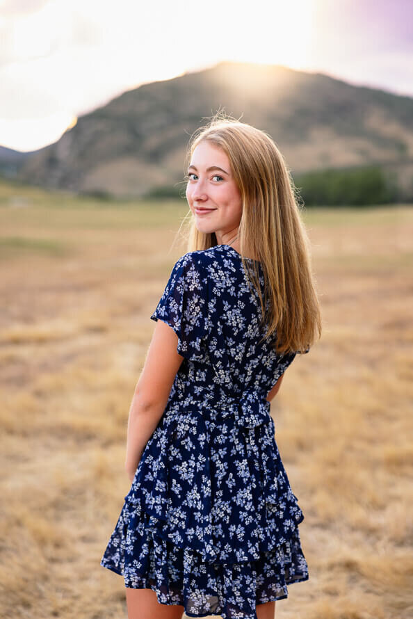 Senior girl in blue dress looks over her shoulder with mountains in the background.