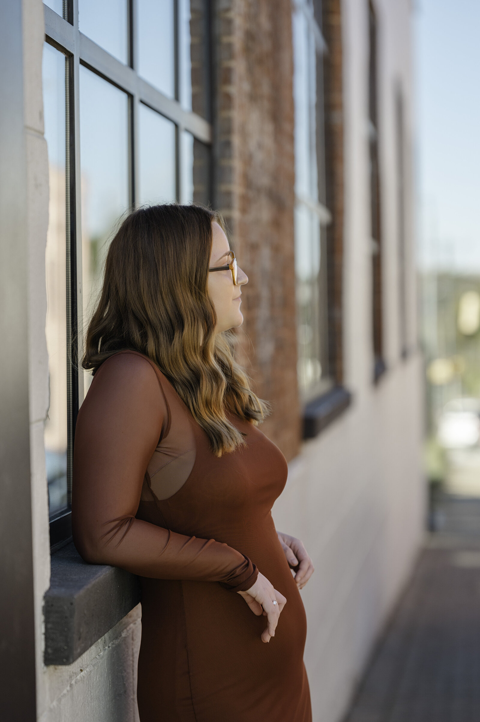 Senior leans against brick building while wearing a rust color dress  and looking into the distance