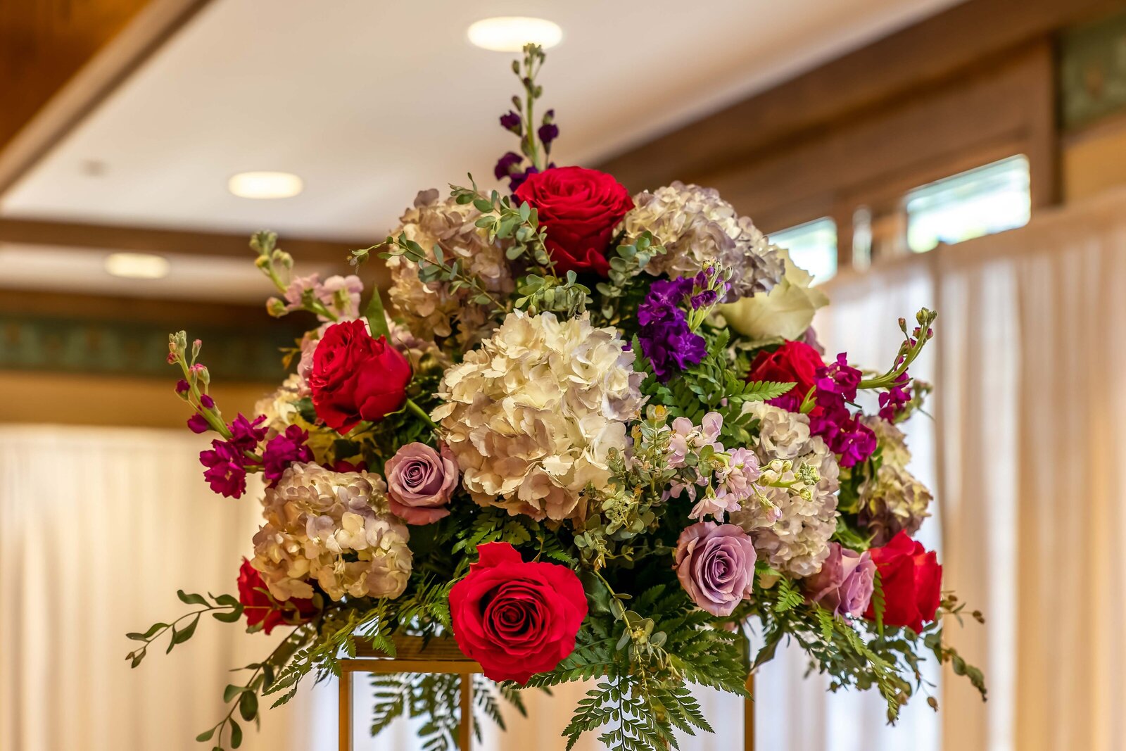An elevated wedding centerpiece with vibrant red roses, white hydrangeas, and purple accents, captured at a Chetola Resort, Blowing Rock, NC wedding reception, blending bold elegance and natural beauty.